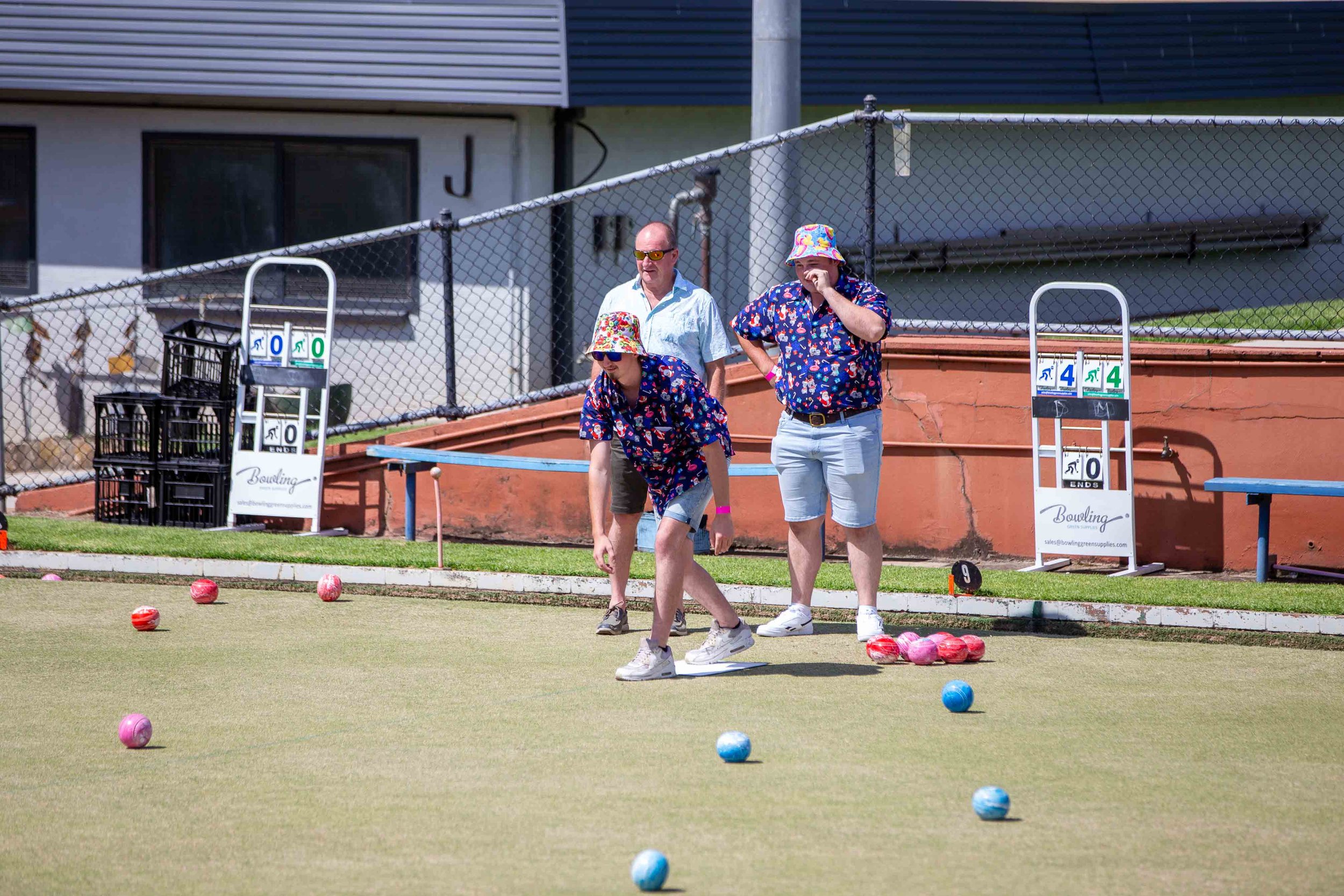 Three people playing lawn bowls on a green, with a fence and residential building in the background. Two women and one man, dressed in colorful summer clothing and hats, are aiming at bowls scattered across the grass.