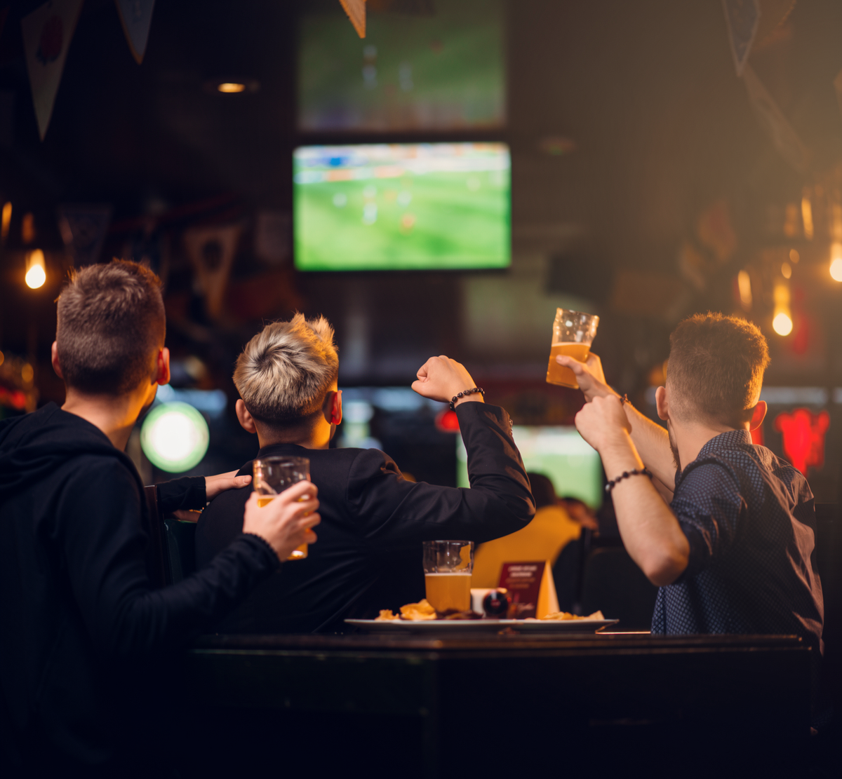 Group of young men watching a soccer game on television in a bar, some raising glasses of beer in celebration.