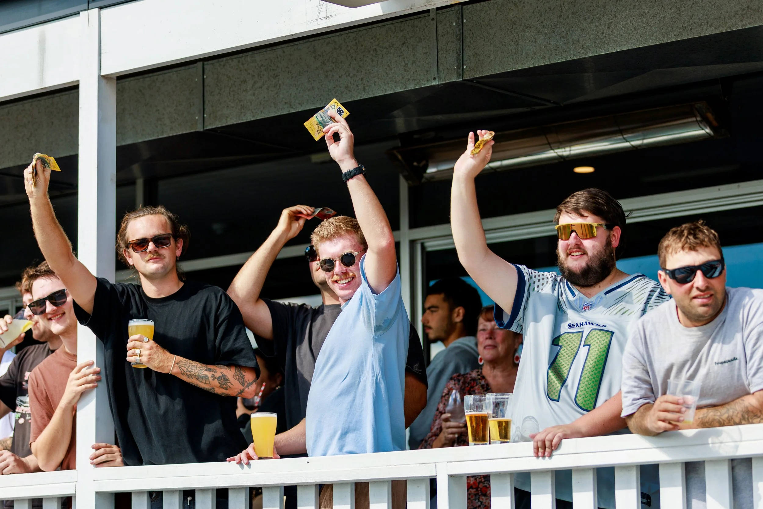 Group of people celebrating outside, with some raising glasses and holding snacks. Most are wearing casual clothes and sunglasses, standing on a balcony or terrace.