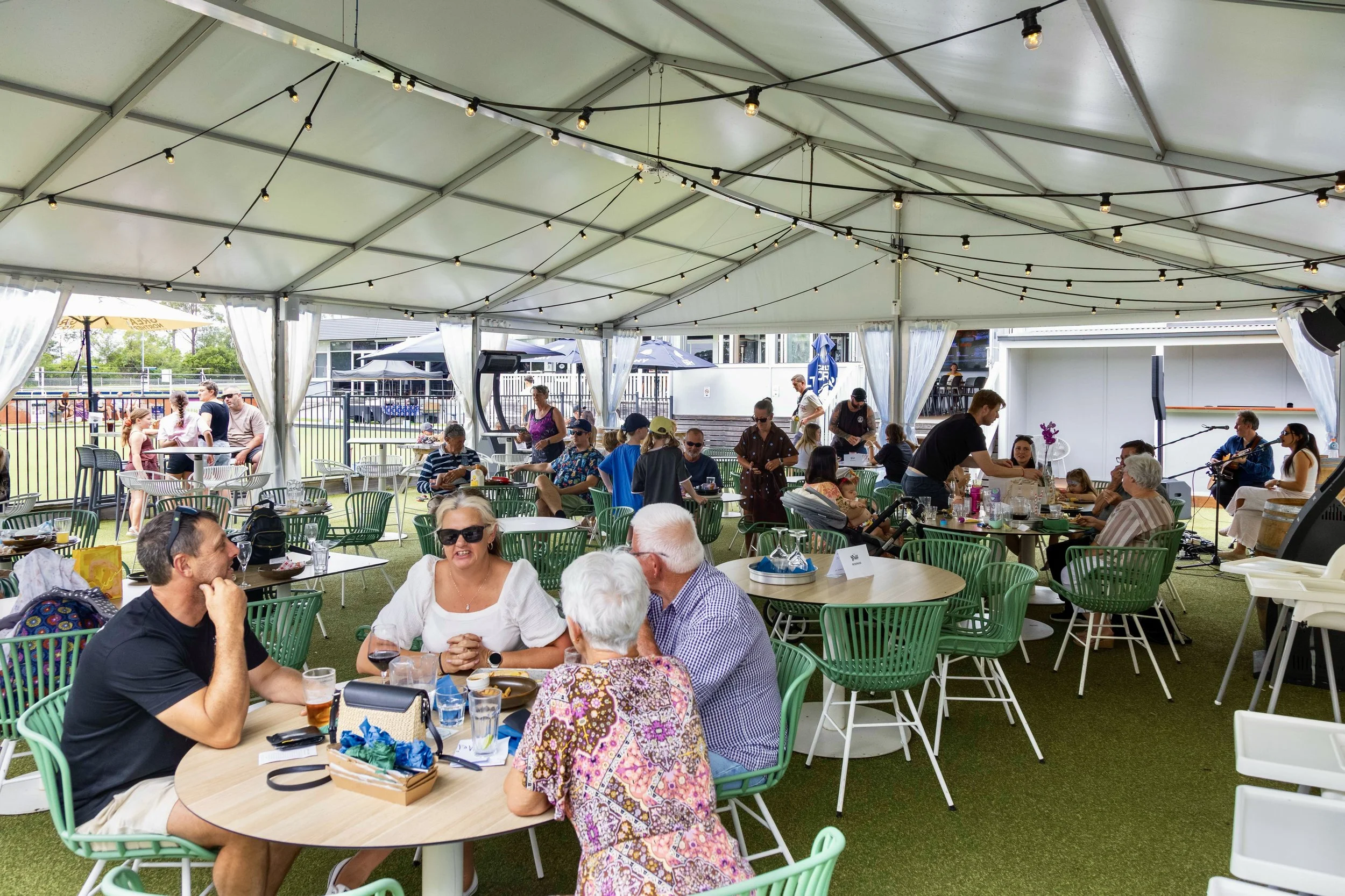 People dining under a large white event tent with string lights, at an outdoor gathering or celebration.