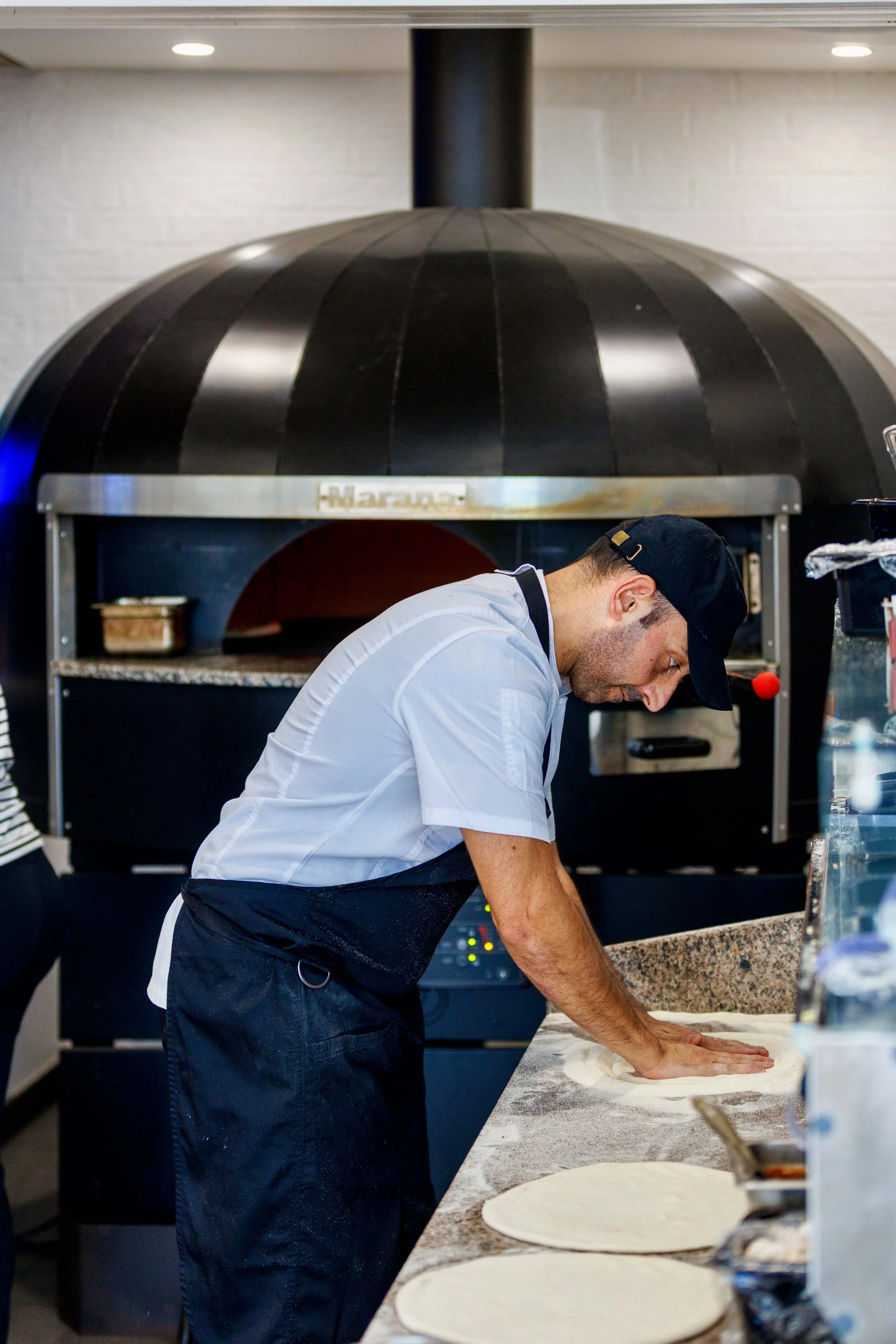 A chef in a white uniform and black apron rolling out pizza dough on a marble countertop in a restaurant kitchen, with a large black pizza oven in the background.
