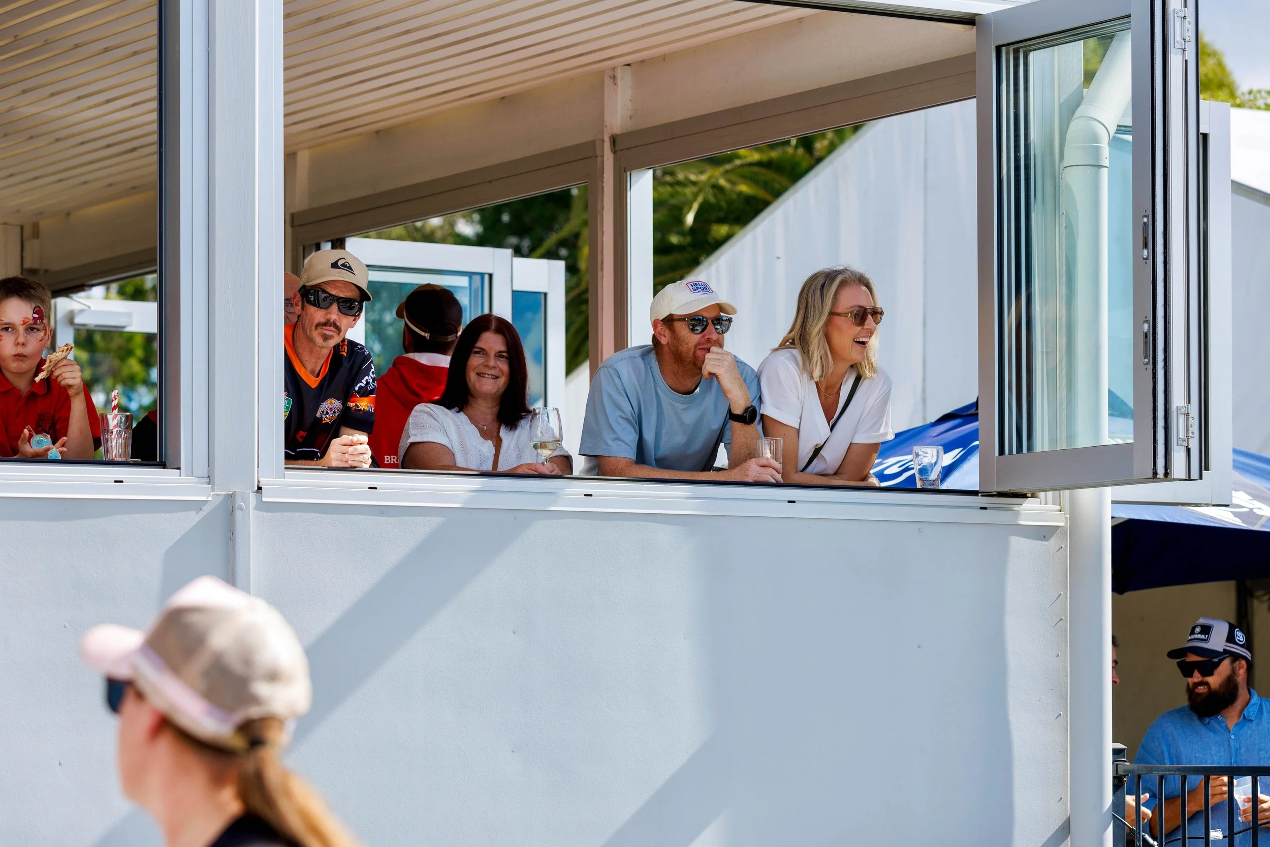 People watching an outdoor event from a balcony, including a woman with sunglasses, a man with a cap, and a young girl, with some individuals below and others outside.