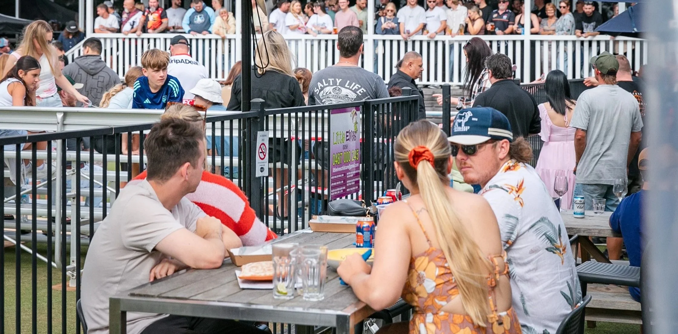 People sitting at an outdoor event, with a group in the foreground at a table and others standing or sitting behind a black fence, watching something in the background.