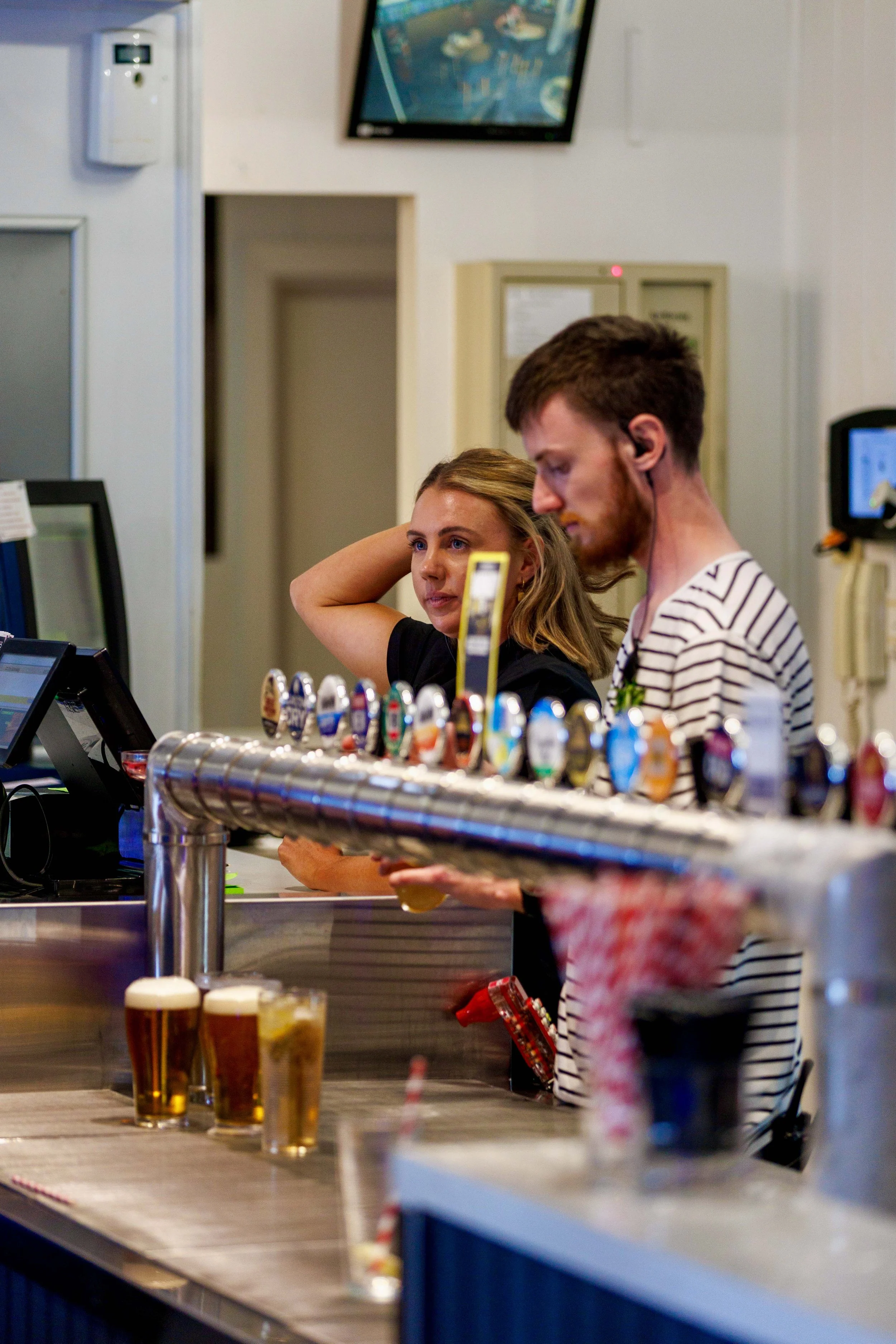 Two people, a woman and a man, standing at a bar counter in a bar or brewery, with glasses of beer in front of them. The woman has blonde hair and is resting her arm on her head, while the man has brown hair and a beard, and is looking down.