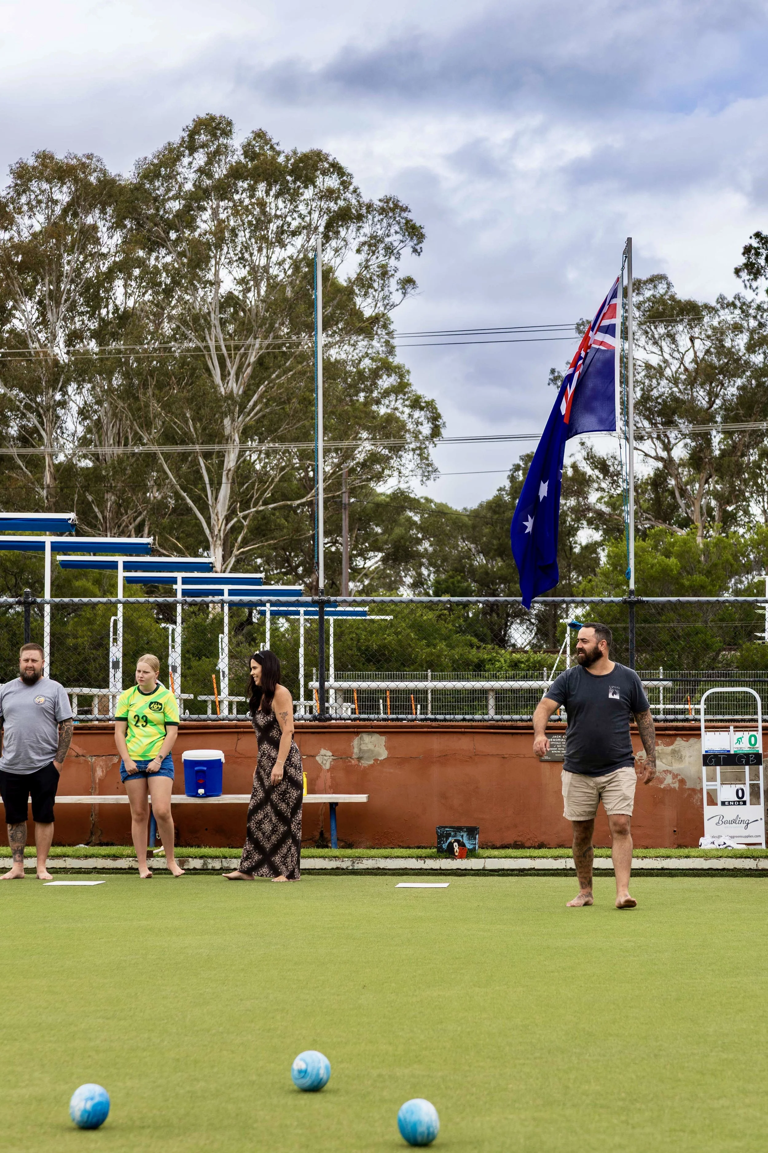 People playing lawn bowls on a green field, with a scoreboard in the background and flags flying above the field.