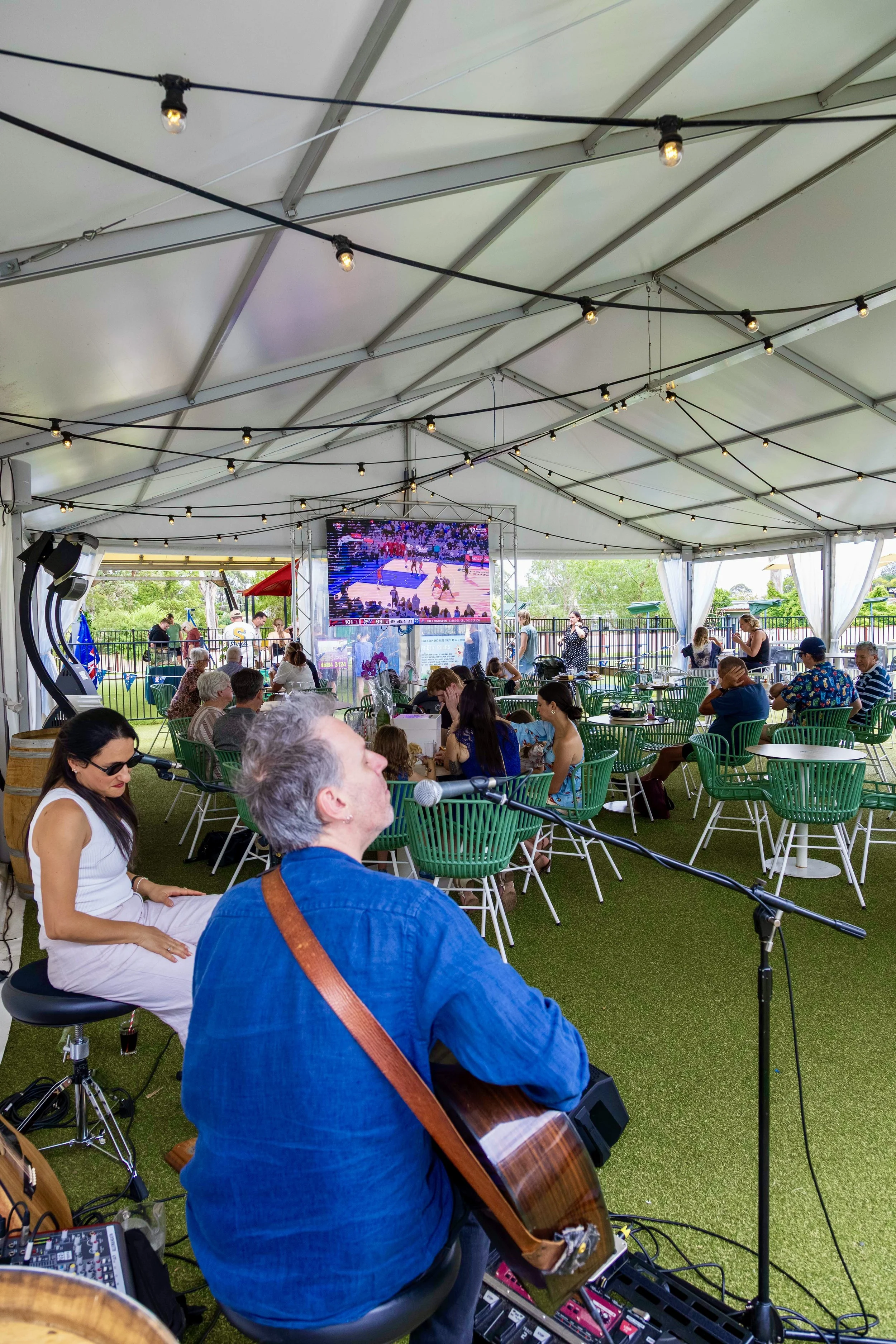 People gathered under a large white tent watching a sports game on a big screen. There are string lights hanging from the ceiling, tables and chairs, and a musician playing guitar in the foreground.