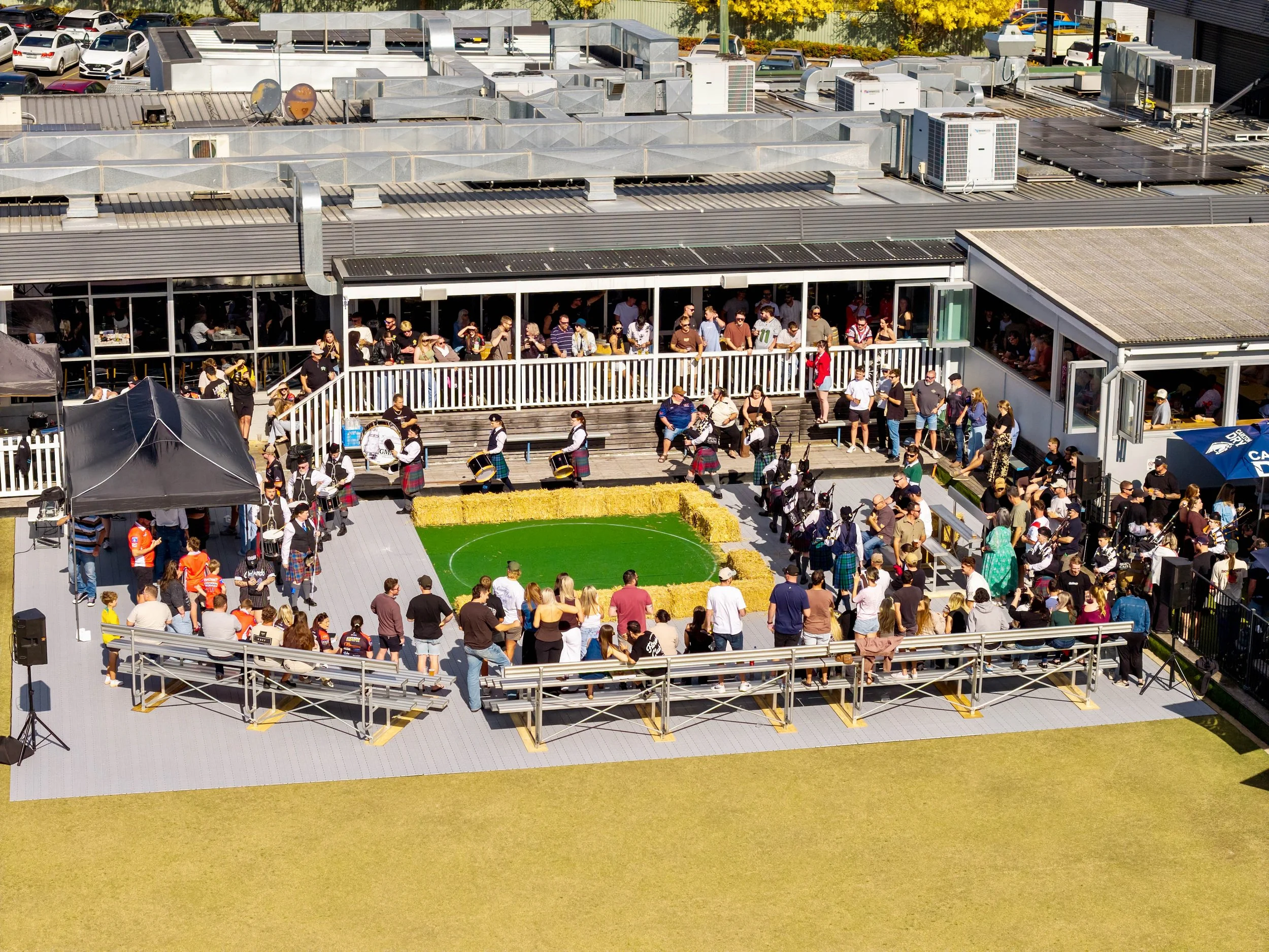 A crowd of people gathered on a rooftop terrace, with some standing and some sitting, and a band playing instruments in the foreground. The terrace has railings and is part of a larger building with parking lot and multiple rooftop HVAC systems visib