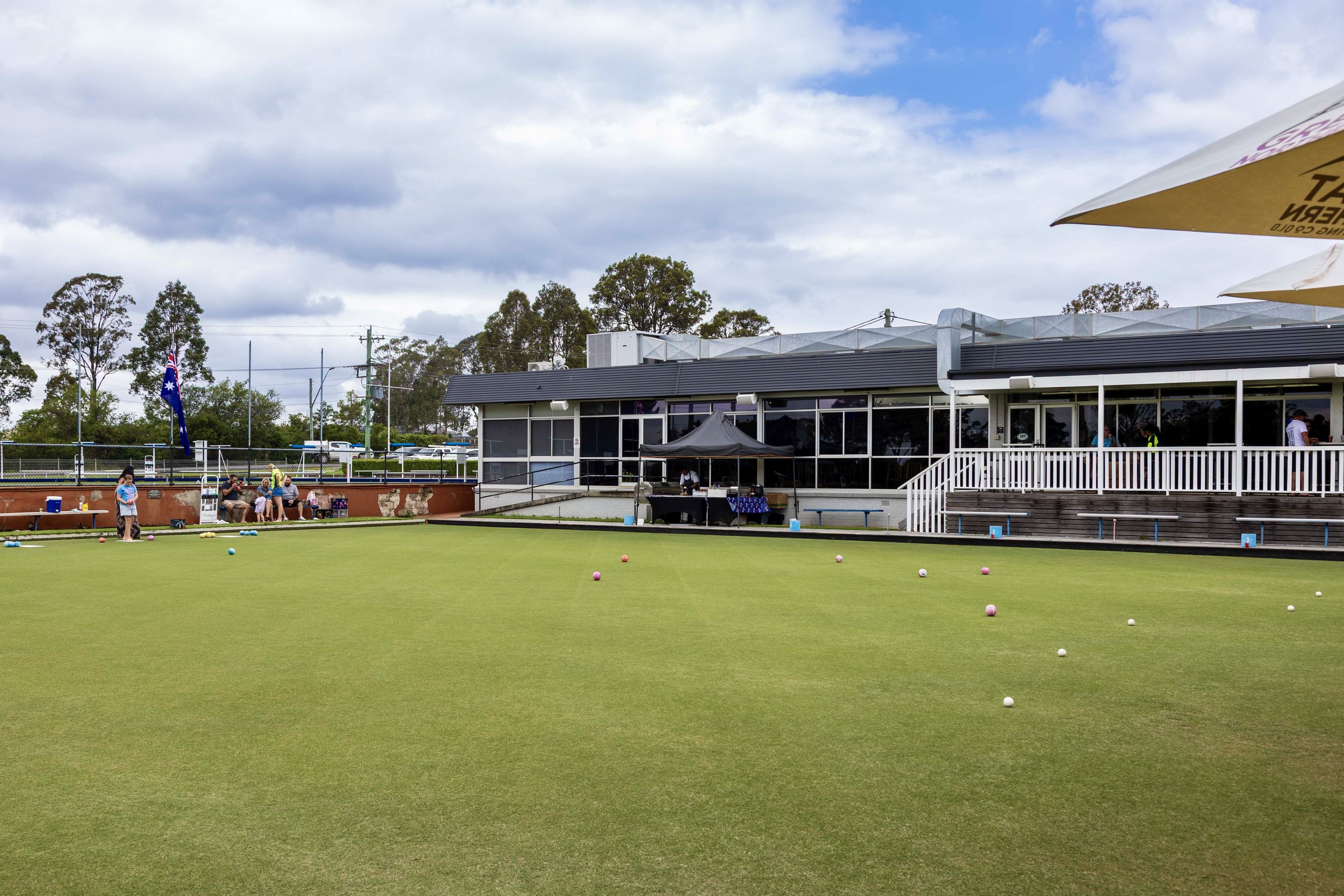 Lawn bowls court with colorful bowls scattered on the green, a building with large windows and patio area, and people sitting and standing nearby. There are trees, a flag, and outdoor umbrellas in the background.