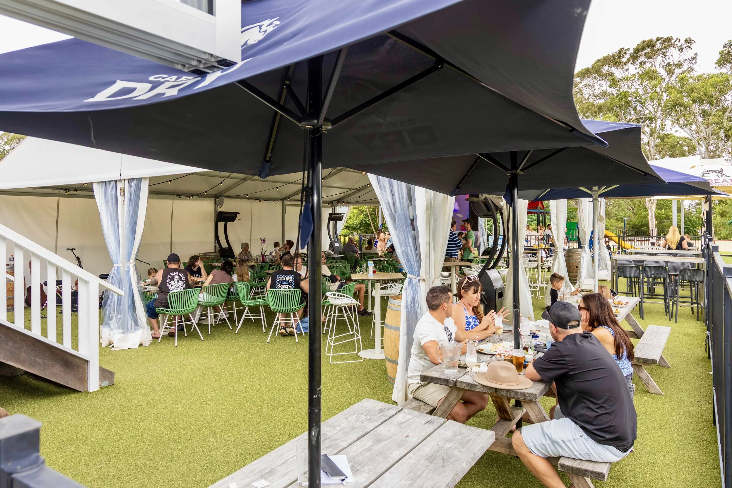 Outdoor dining area with people sitting at tables under umbrellas and a tent, with trees in the background.