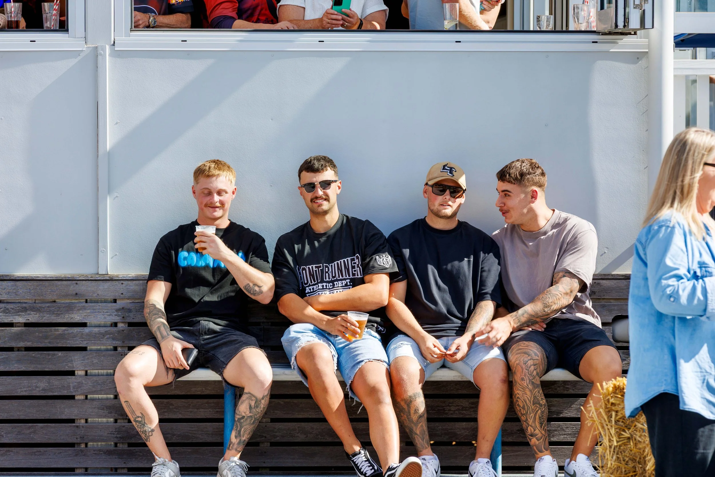 A group of five young men sitting on a wooden bench outside, casually dressed, some holding drinks, with a white wall behind them. Two are wearing sunglasses and a tan cap, another is holding a phone, and they appear to be socializing on a sunny day.
