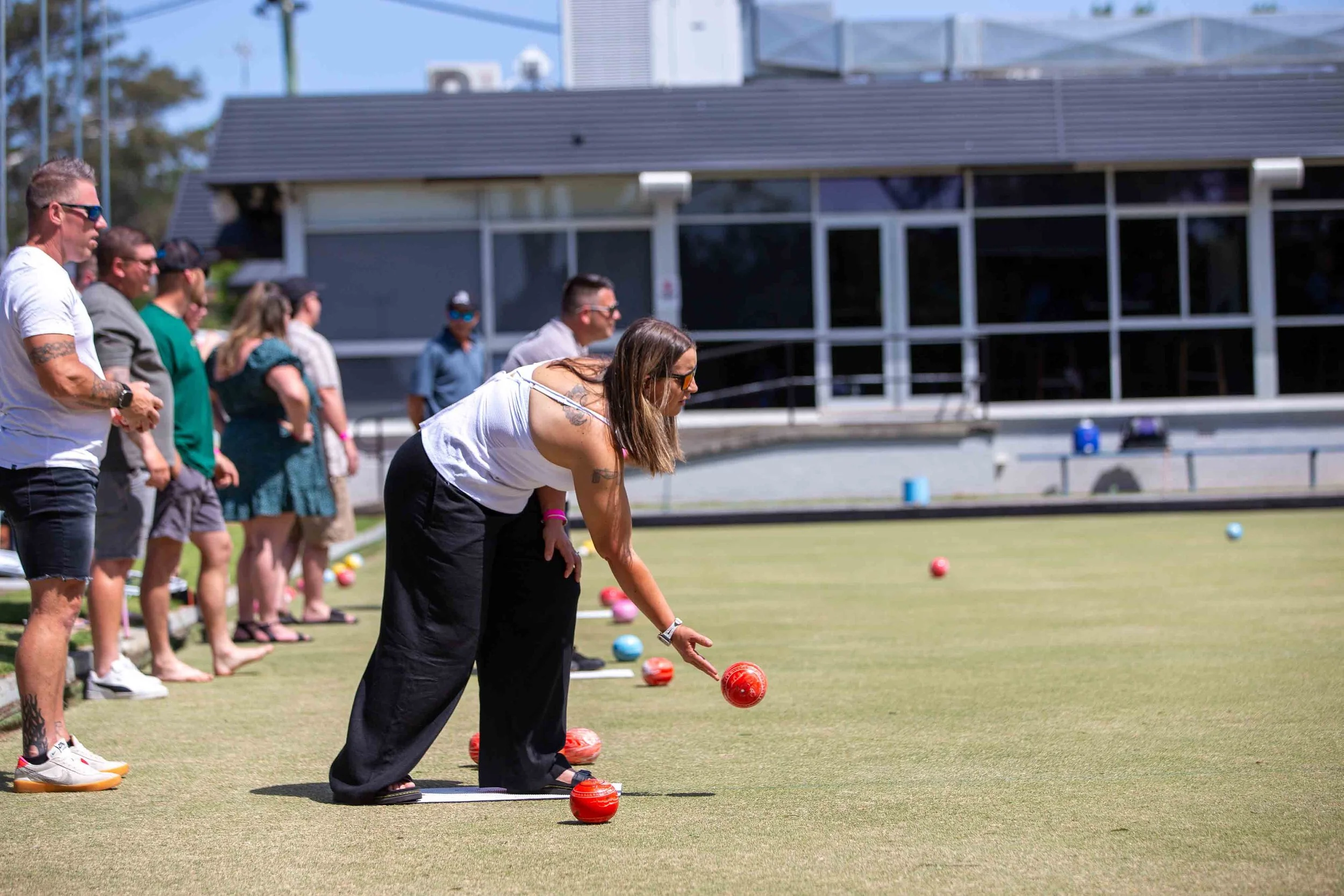 A group of people, mostly women, are lined up on a lawn playing lawn bowls, with one woman in the foreground aiming before rolling a red ball. The scene is outdoors on a sunny day, with a building in the background.