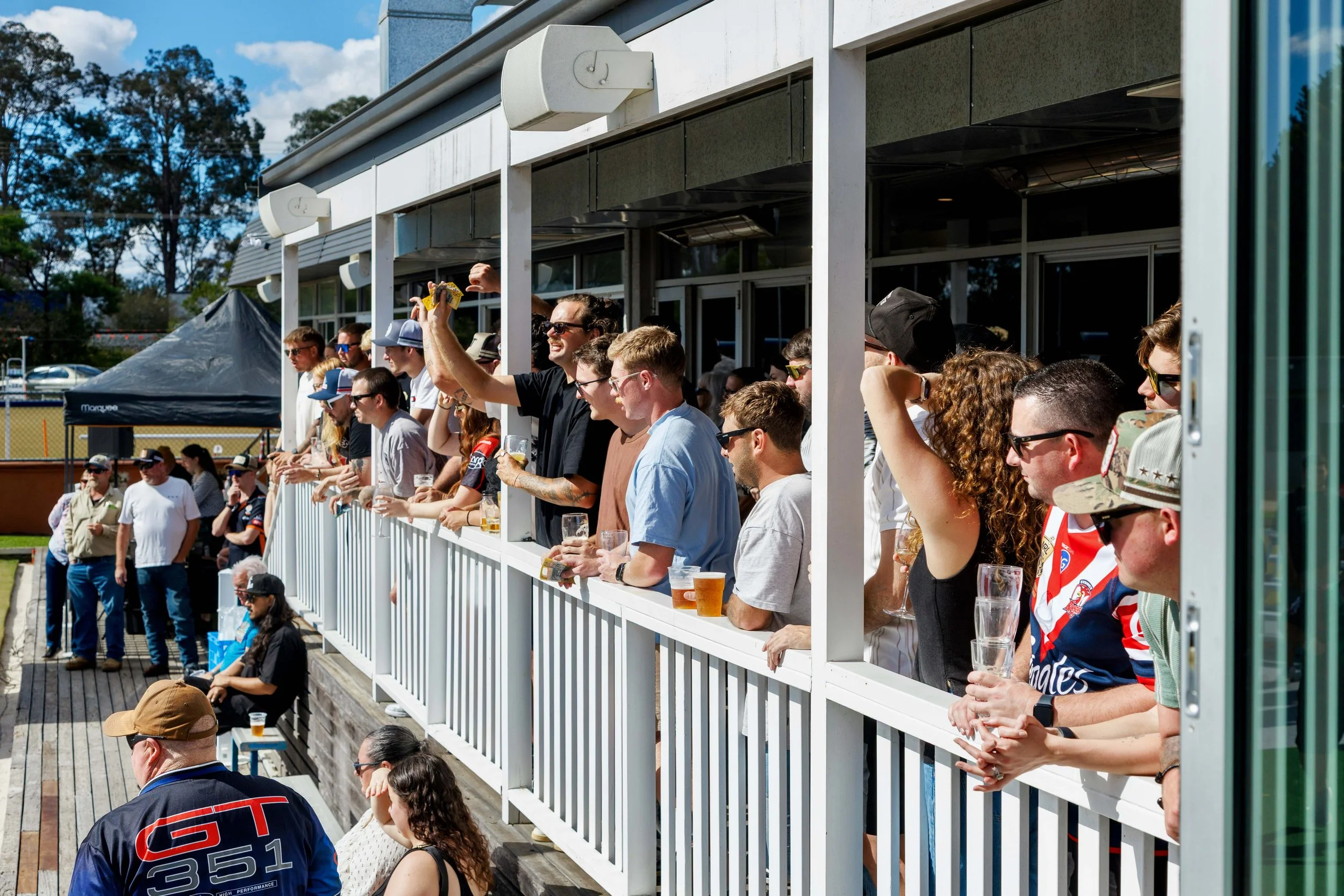 People standing on a balcony, watching an outdoor event, some holding drinks, under a blue sky with scattered clouds.
