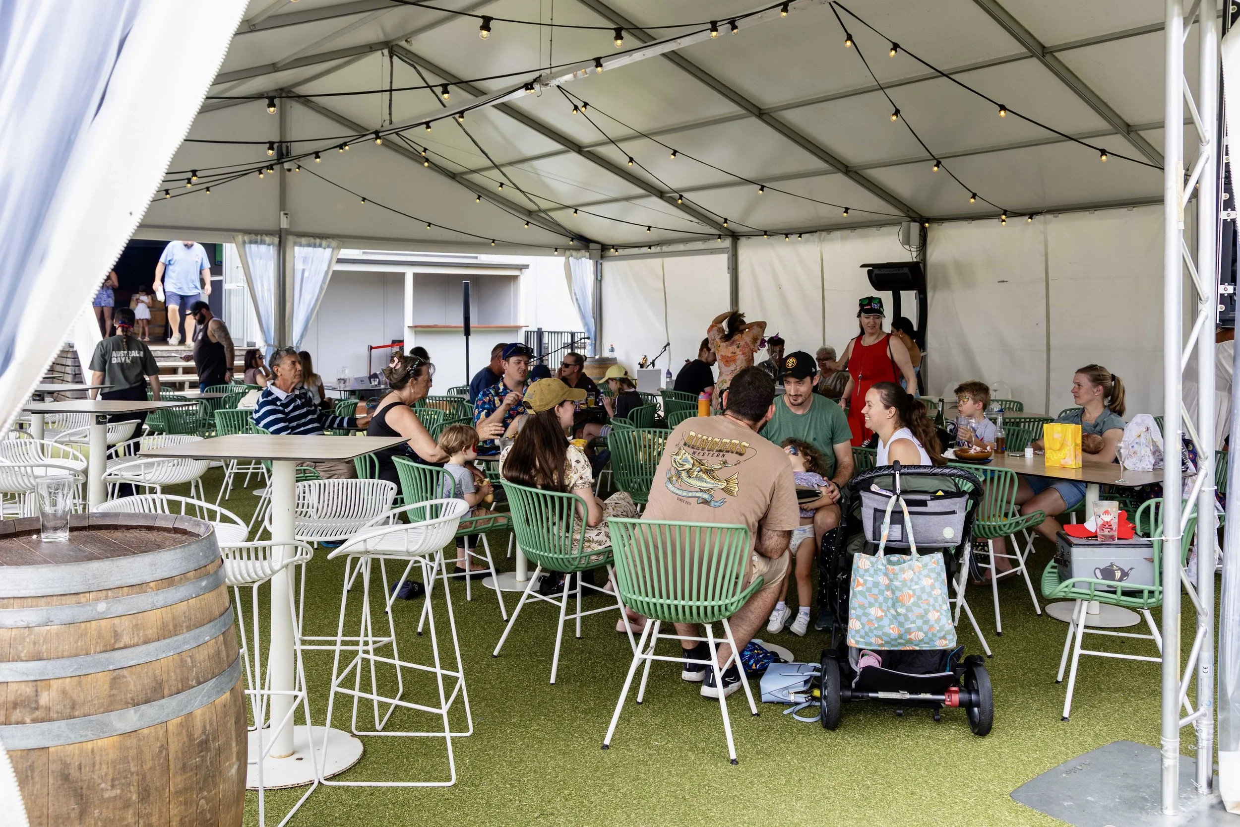People sitting and dining under a large white tent with string lights, with some children and adults, at what appears to be a casual outdoor event or festival.