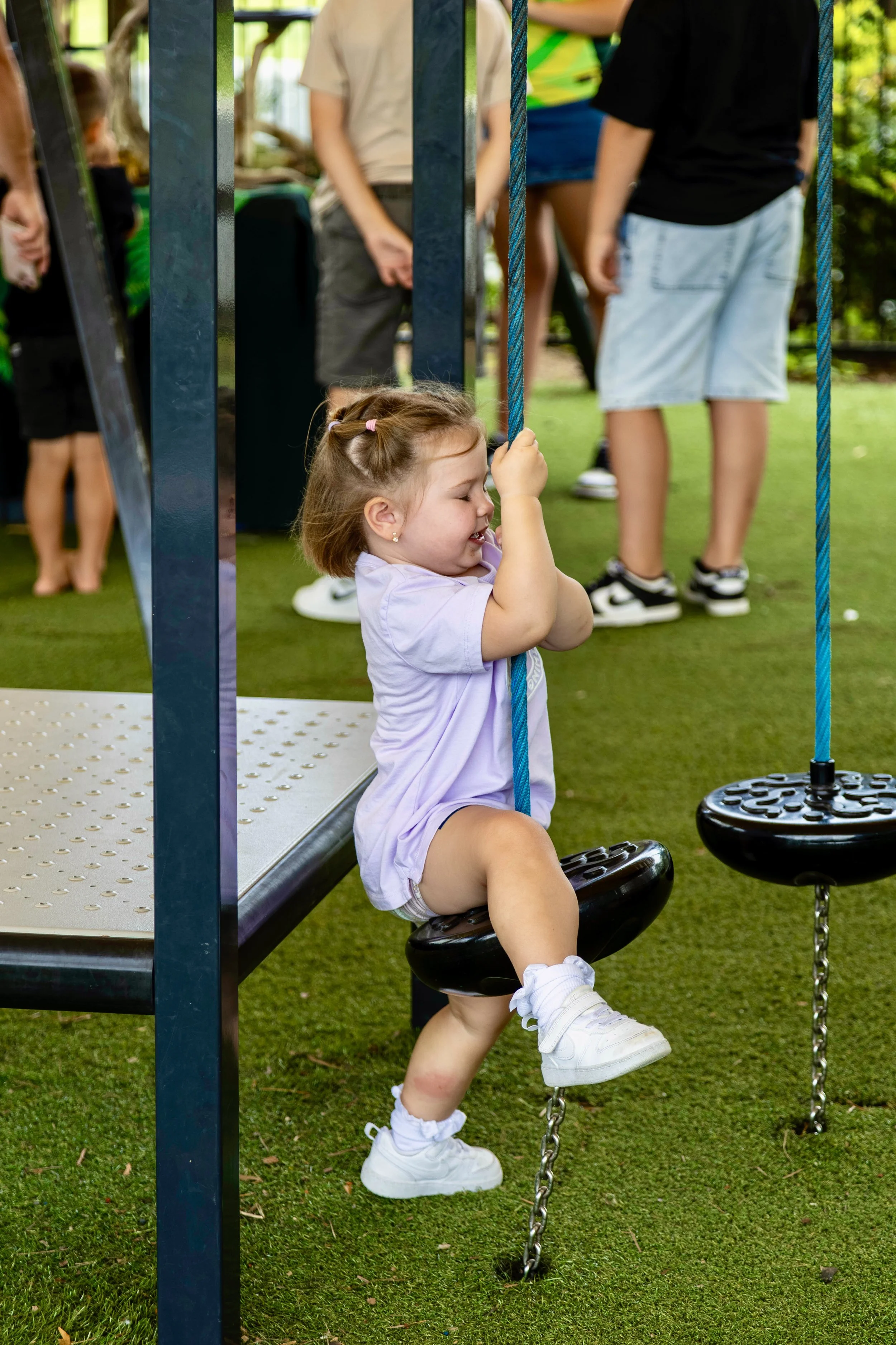 A young girl sitting on a playground swing, holding the chains and smiling while in a purple outfit and white sneakers. Other children are visible in the background, along with green grass and outdoor trees.