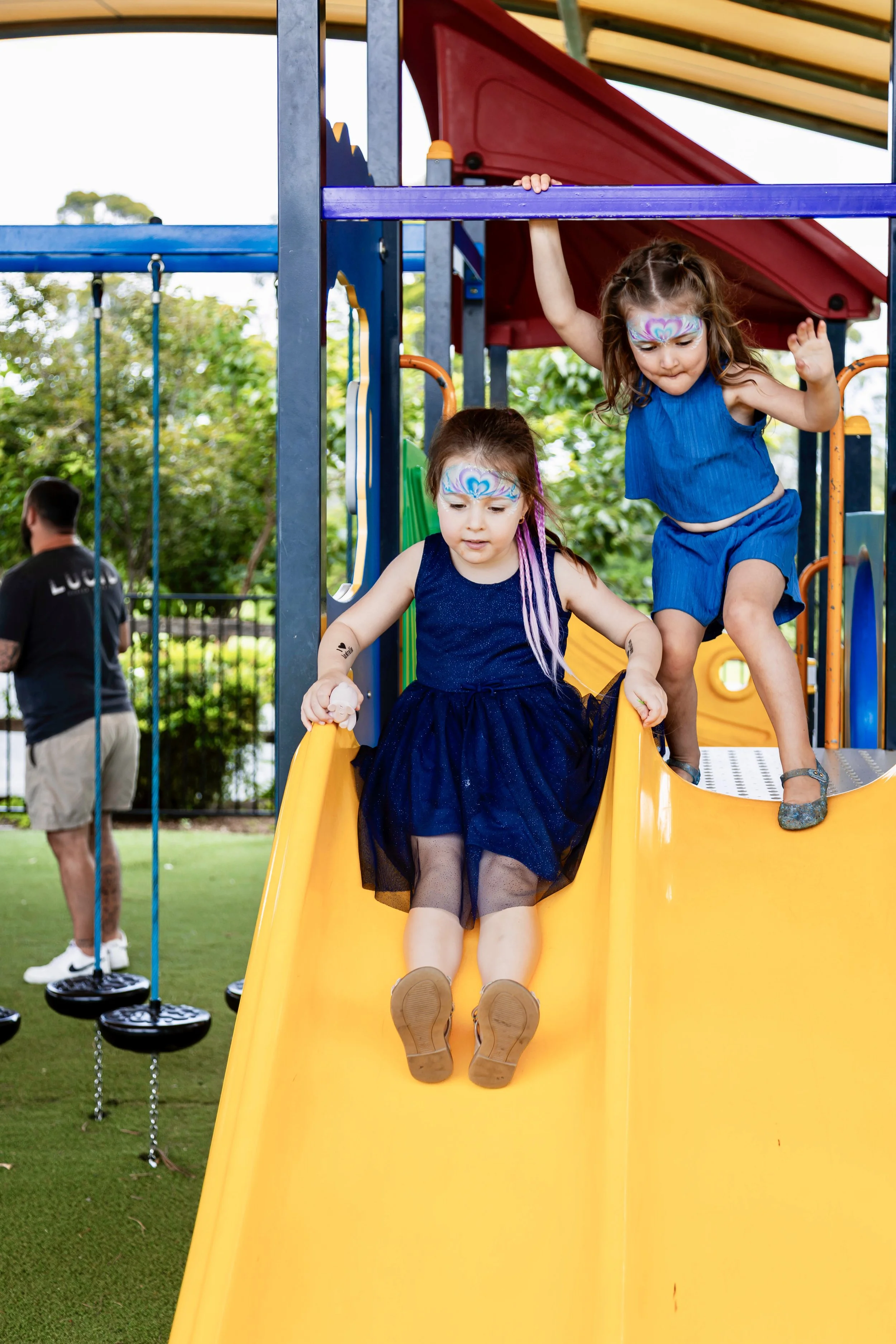 Two young girls with face paint on their faces go down a yellow slide at a playground. One girl in a blue dress is at the top of the slide, while the other in a dark dress is at the bottom, about to slide down. The playground is outdoors with trees a
