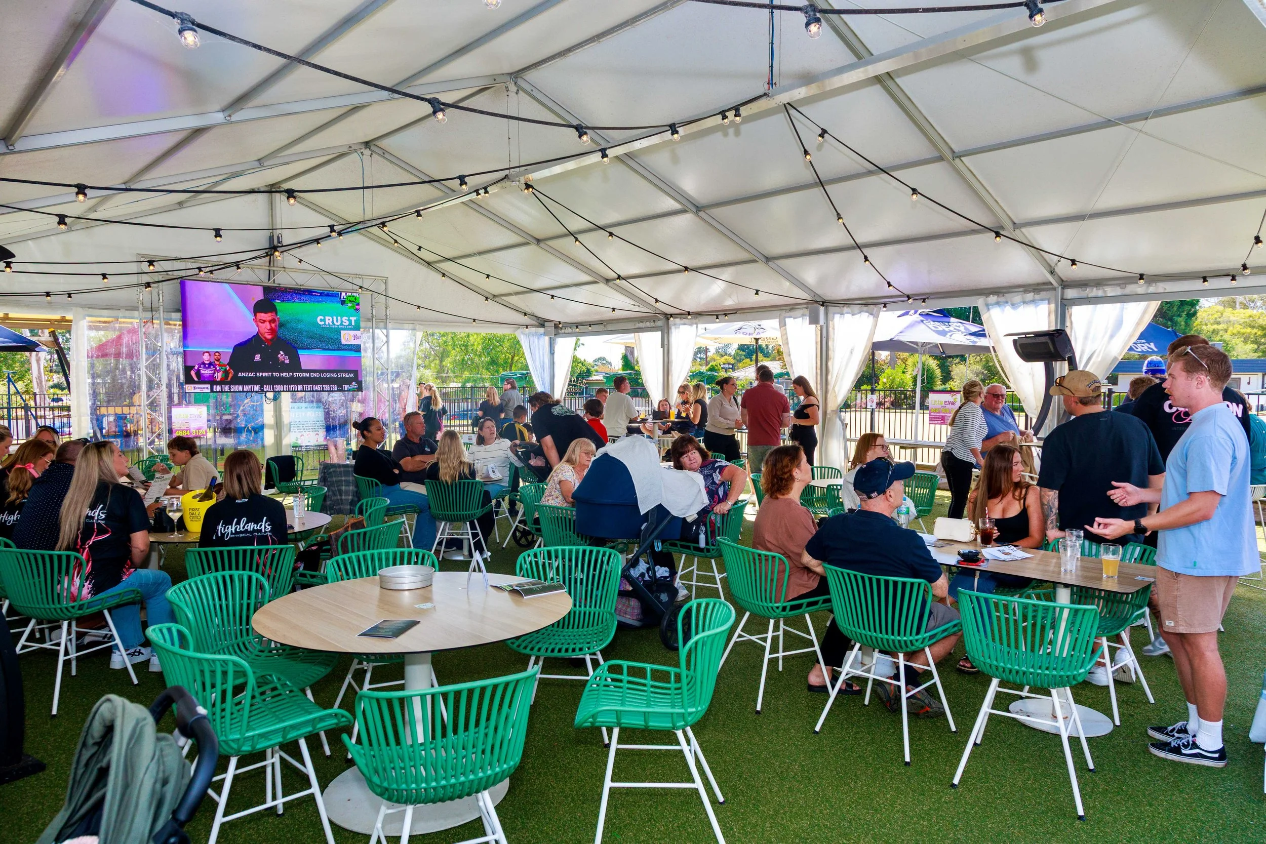 People gathered at an outdoor event under a large tent with string lights, sitting at round tables with green chairs. There is a TV screen displaying a cricket player and a weather forecast. Some individuals are standing and talking, while others are