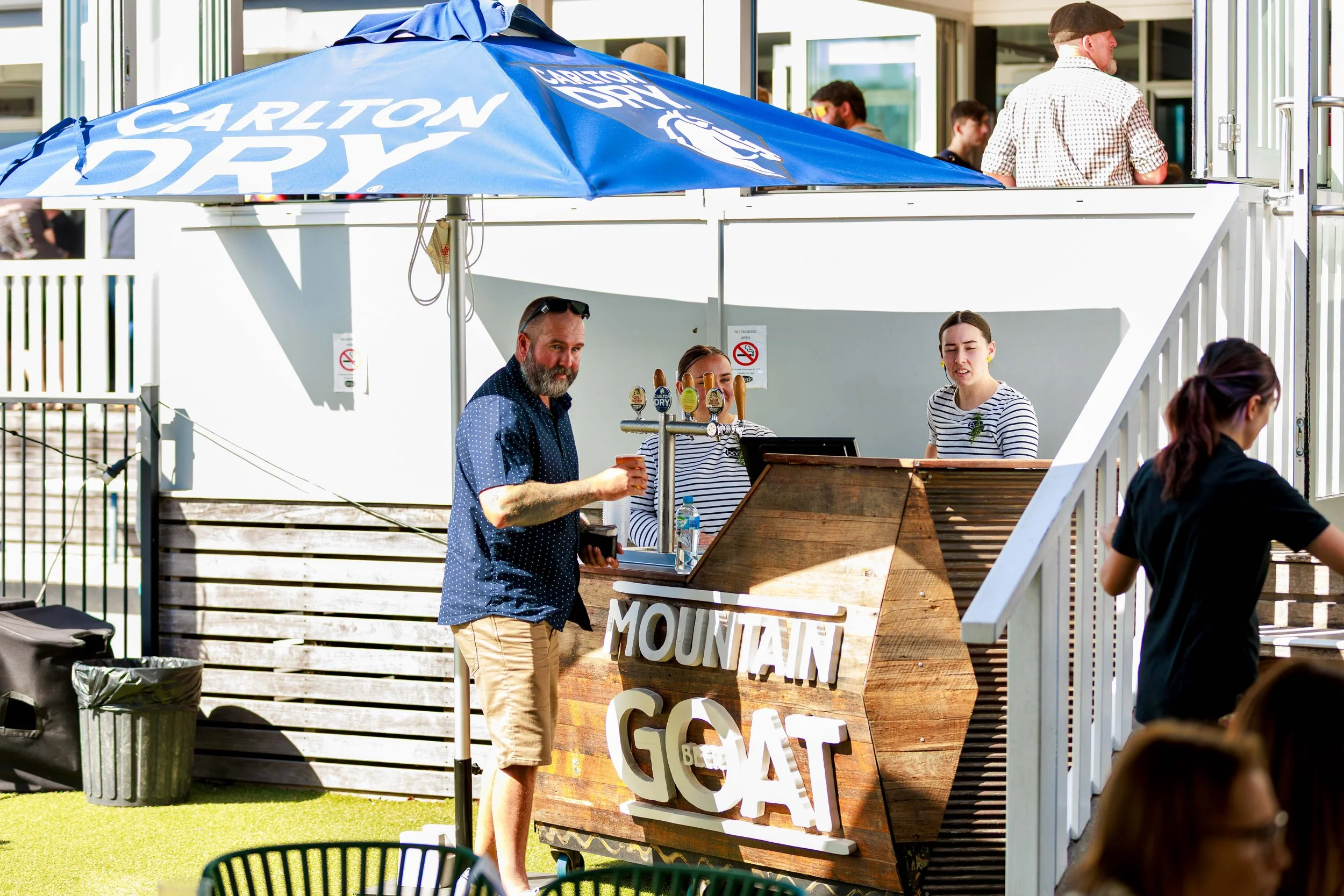 People standing and chatting outside at a beach bar with a large Carlton Dry umbrella, a man at a drink stand, and a wooden sign with partially visible text.