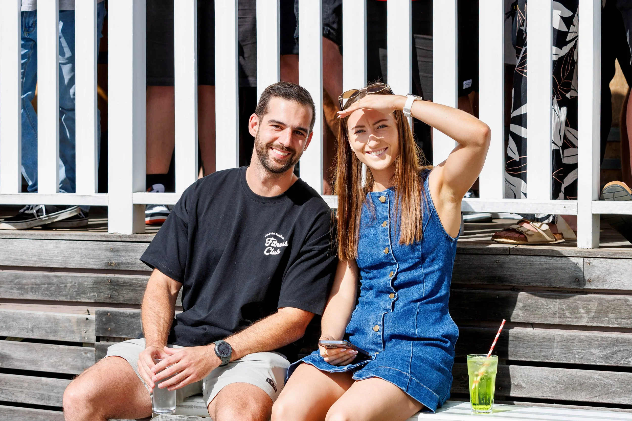 A young man and woman sitting on outdoor wooden steps, smiling at the camera. The woman is shielding her eyes from the sun with her hand and has sunglasses on her head. They have drinks on a table nearby, including a green beverage with a straw. In t