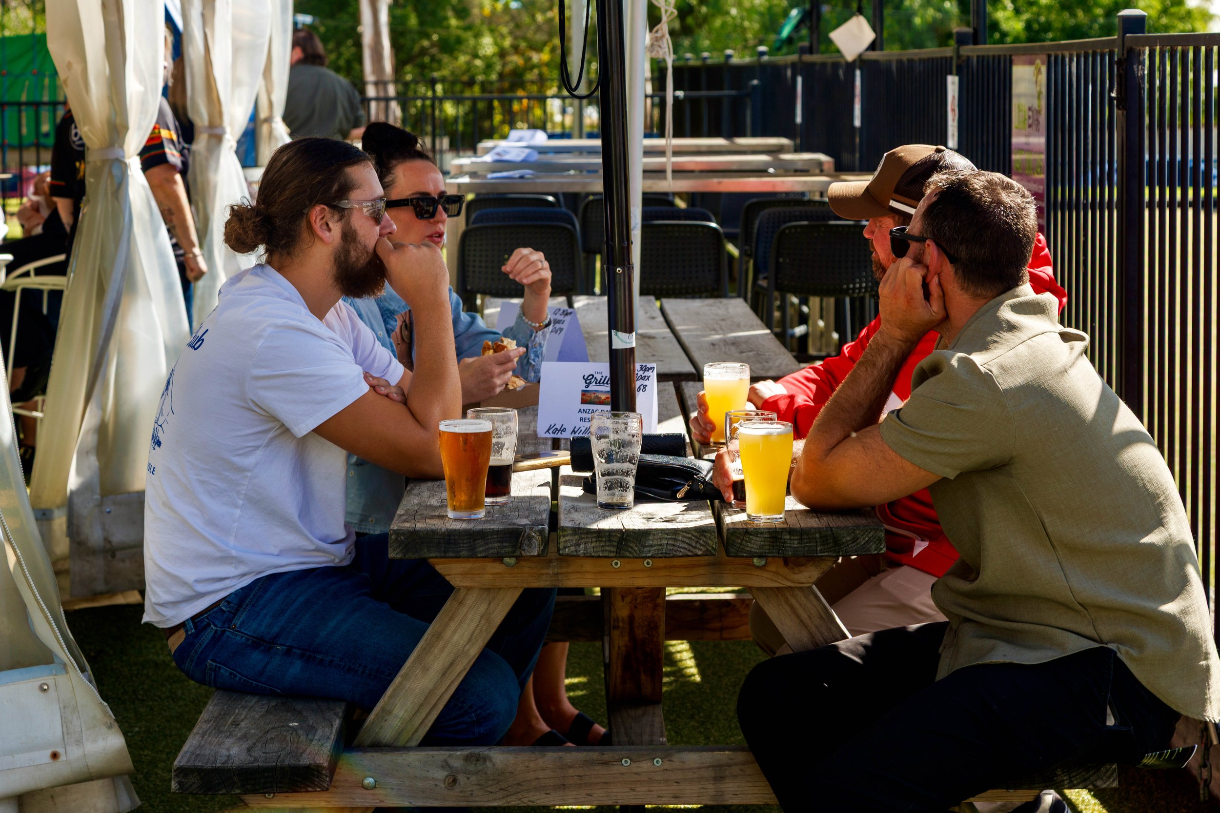 Four people sit around a wooden picnic table outdoors, engaged in conversation, with glasses of beer in front of them. Two men have facial hair and are wearing sunglasses. The woman has her hair up and is wearing sunglasses. The scene takes place in 
