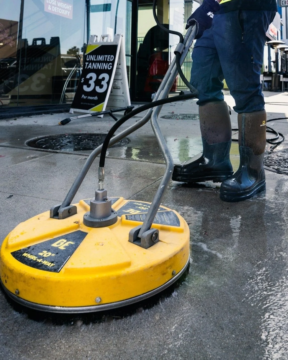 A person operating a wet surface cleaning machine on a sidewalk outside a building with a sign advertising unlimited tanning for $33 per month.