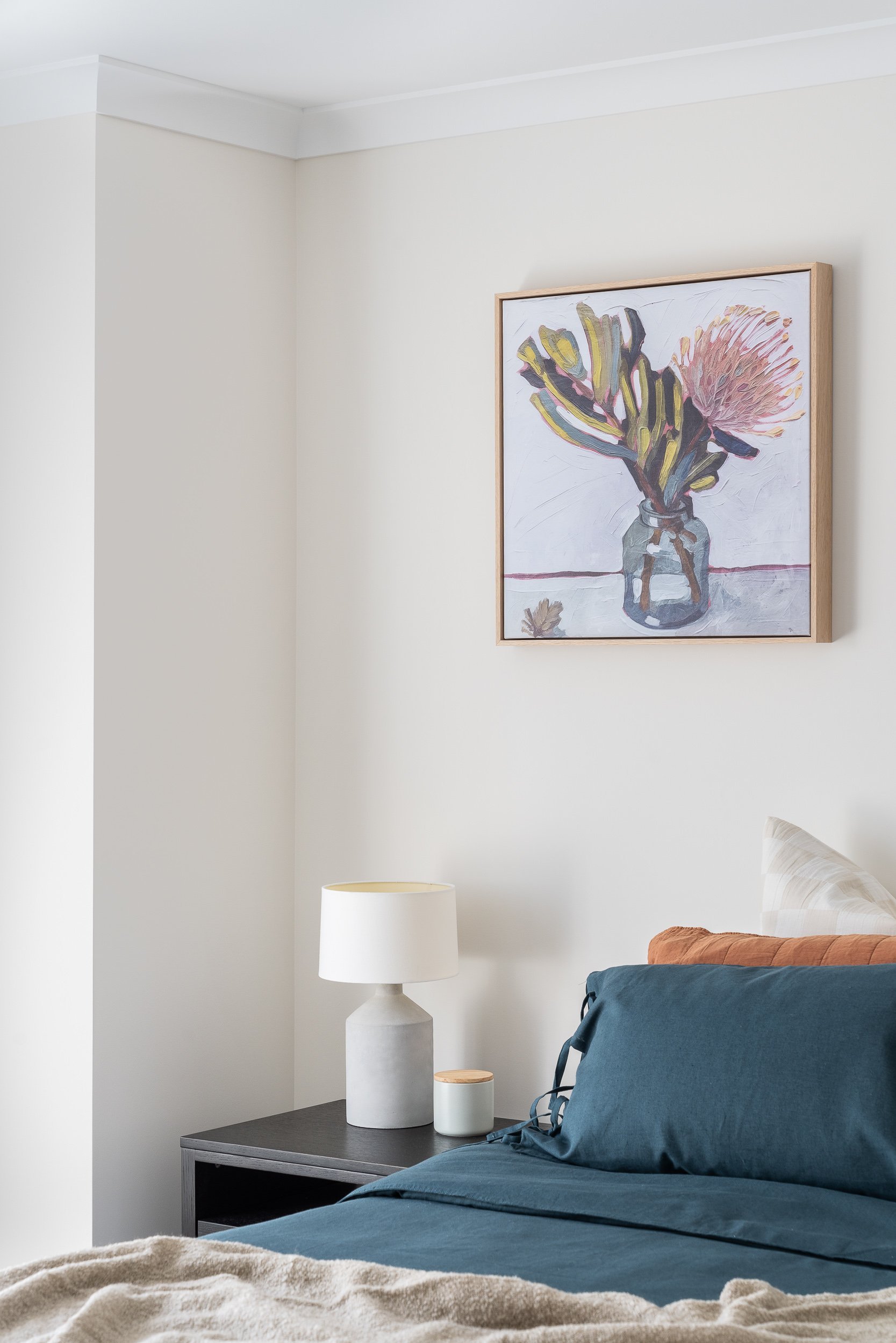 Bedroom corner with a painting of a flower in a vase, a white table lamp on a nightstand, a teal bedding set, and a pillow against a white wall.
