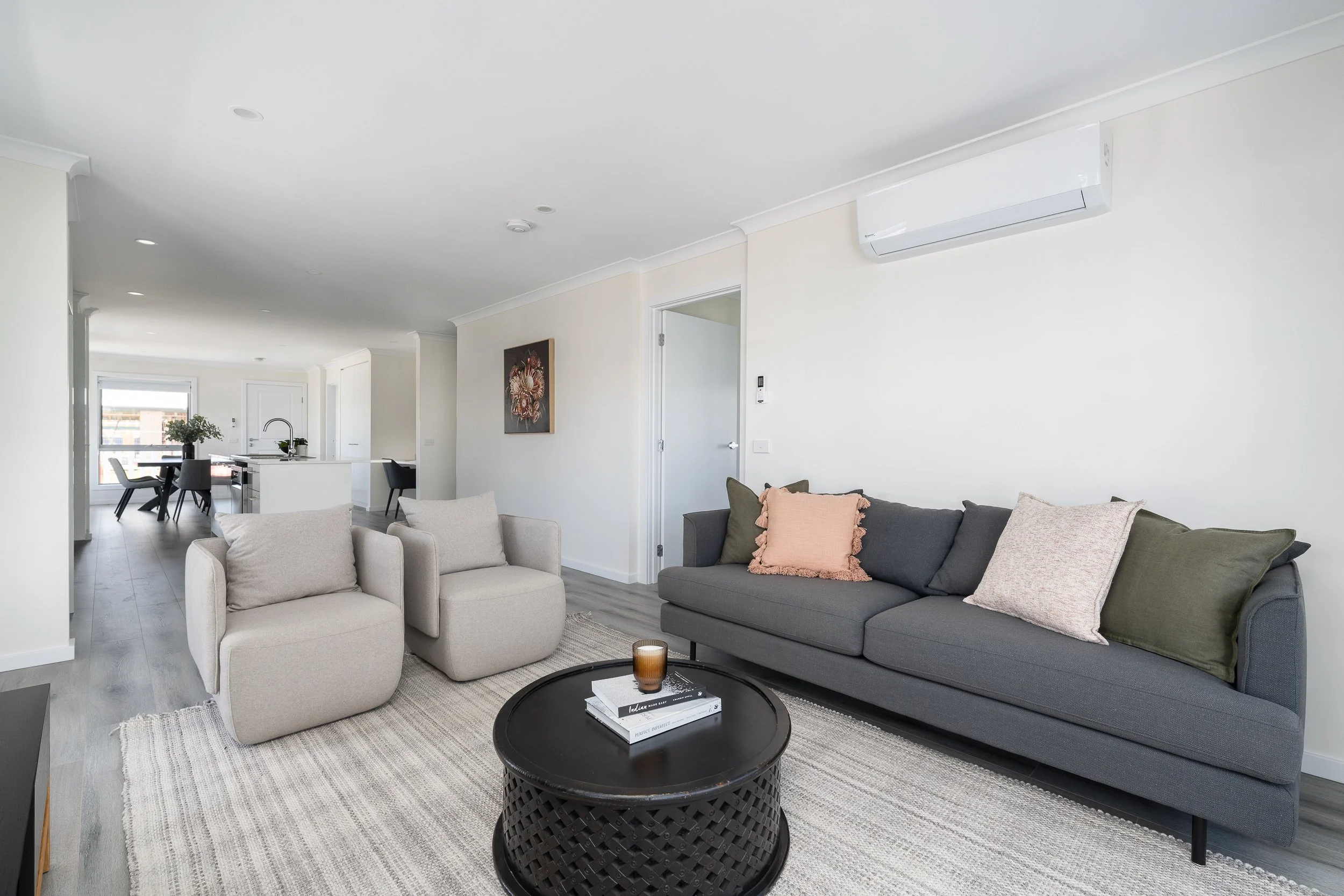 Modern living room with gray sofa, two beige armchairs, black coffee table, and decorative pillows. Open floor plan leads to a kitchen area. Neutral colors and minimalist design.