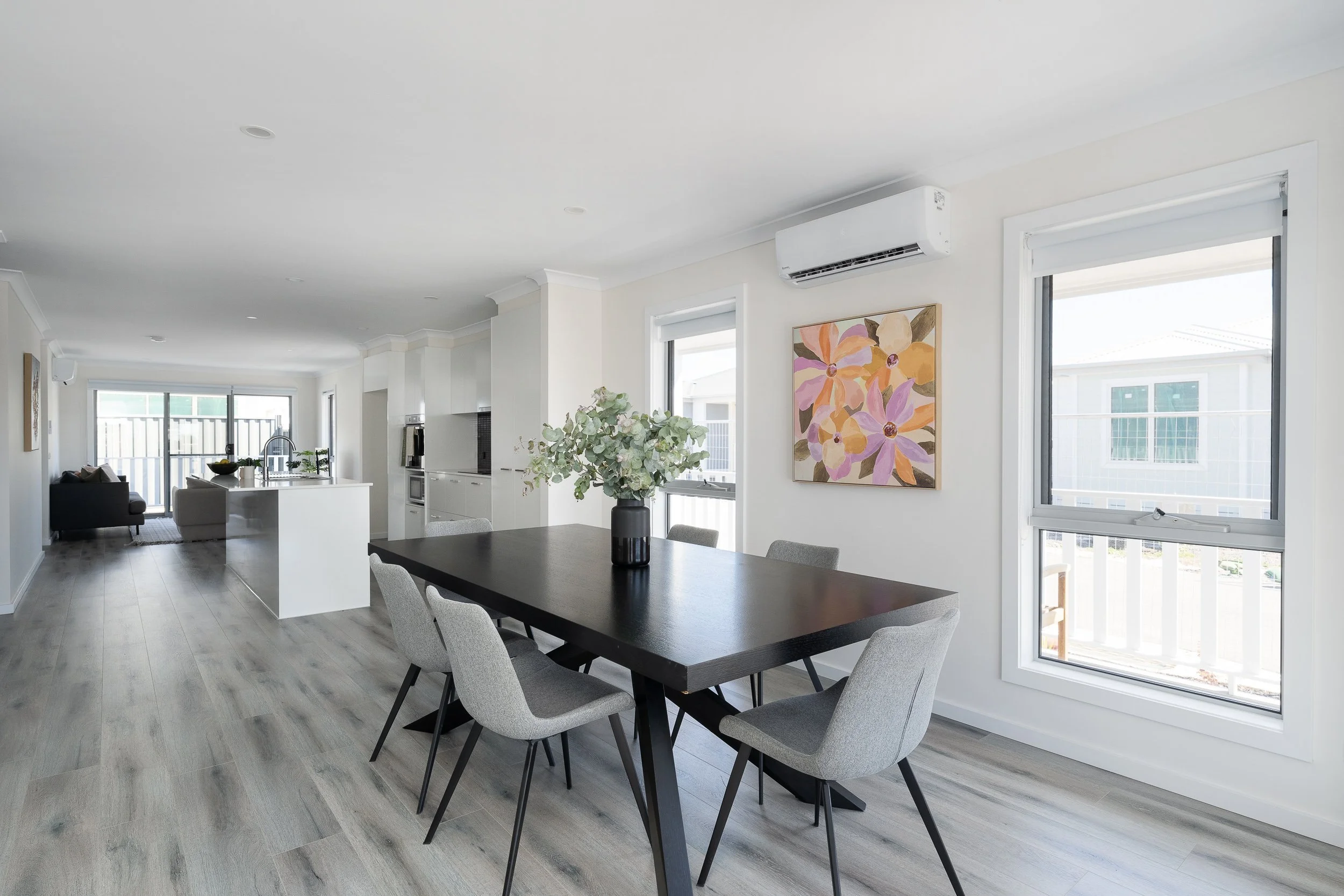 Modern open-plan dining and living area with a black dining table, gray chairs, hardwood flooring, white walls, colorful wall art, and large windows.