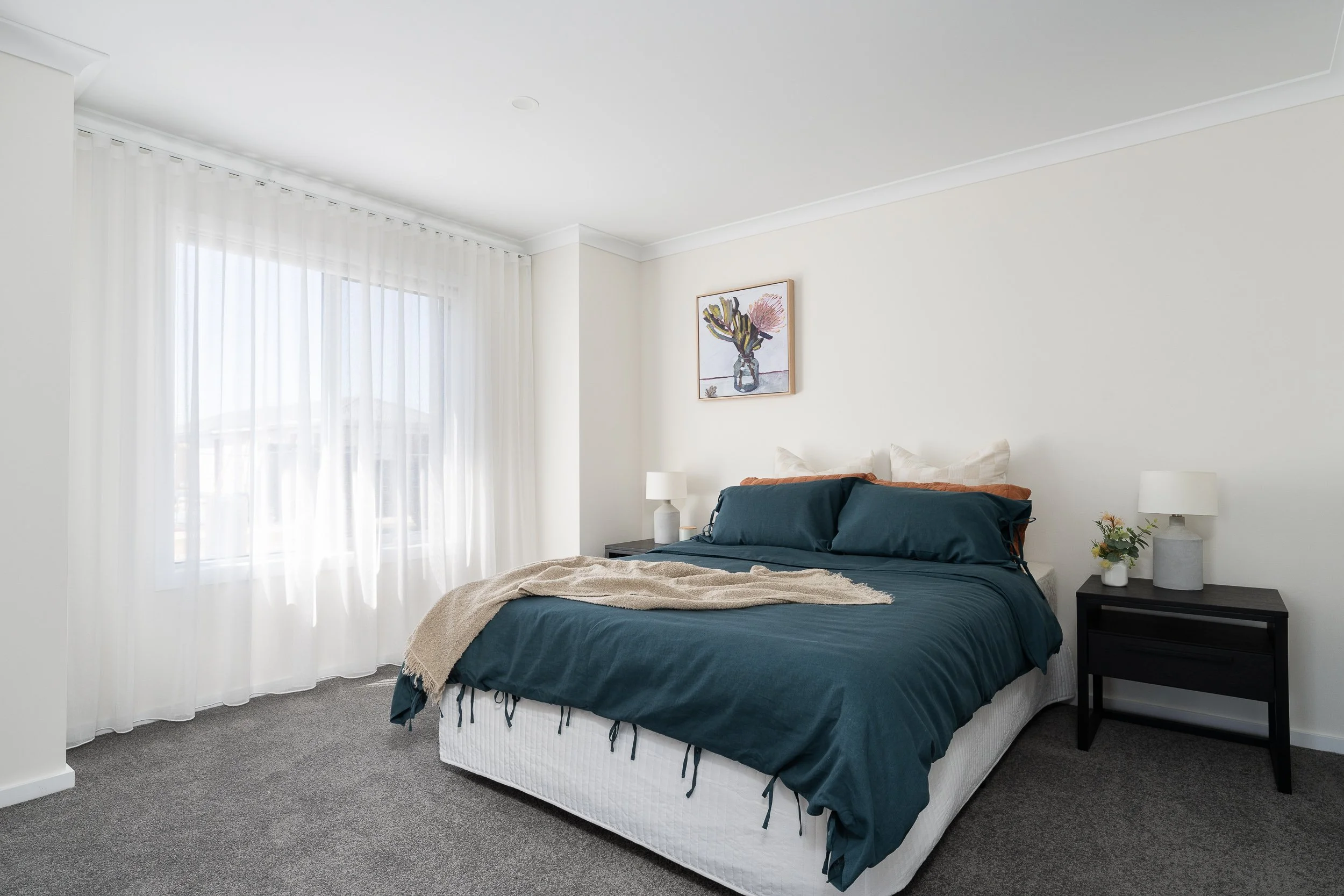 Modern bedroom with a neatly made bed featuring blue bedding, white pillows, and a beige throw blanket. A window with sheer curtains and a framed floral artwork hang above the bed. Two modern nightstands with lamps and a vase of flowers are beside th