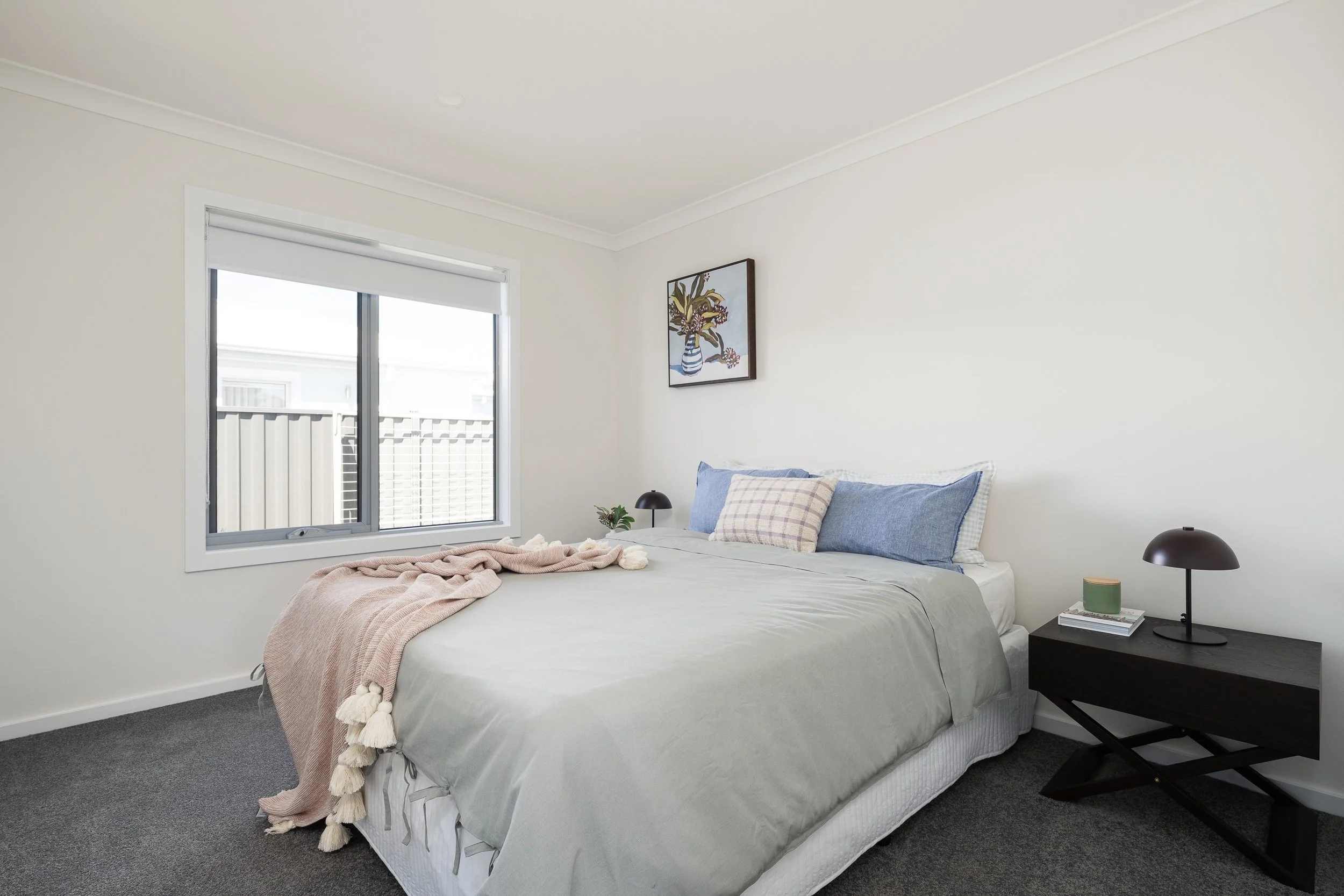 Minimalist bedroom with a bed featuring gray bedding, blue and plaid pillows, and a pink throw blanket. A dark wood nightstand holds a lamp and small decor. A window and framed wall art are in the background.