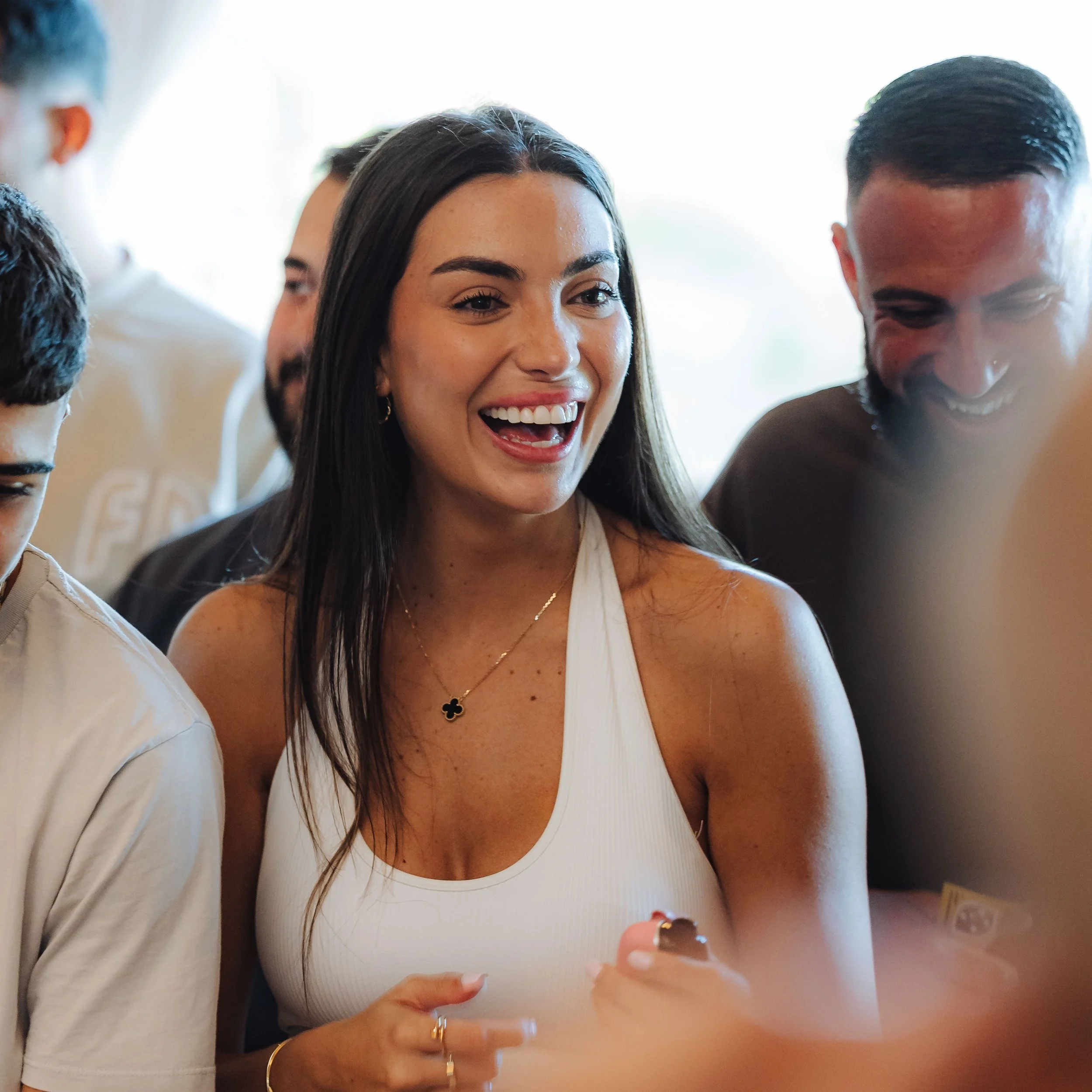 A woman with long dark hair smiling and laughing while holding a phone, surrounded by other people at a social gathering.