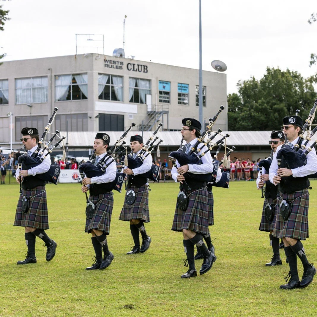 A group of people dressed in traditional Scottish attire, including kilts and bagpipes, performing in a parade or ceremony on a grassy field with onlookers and a building labeled 'West's Rules Club' in the background.