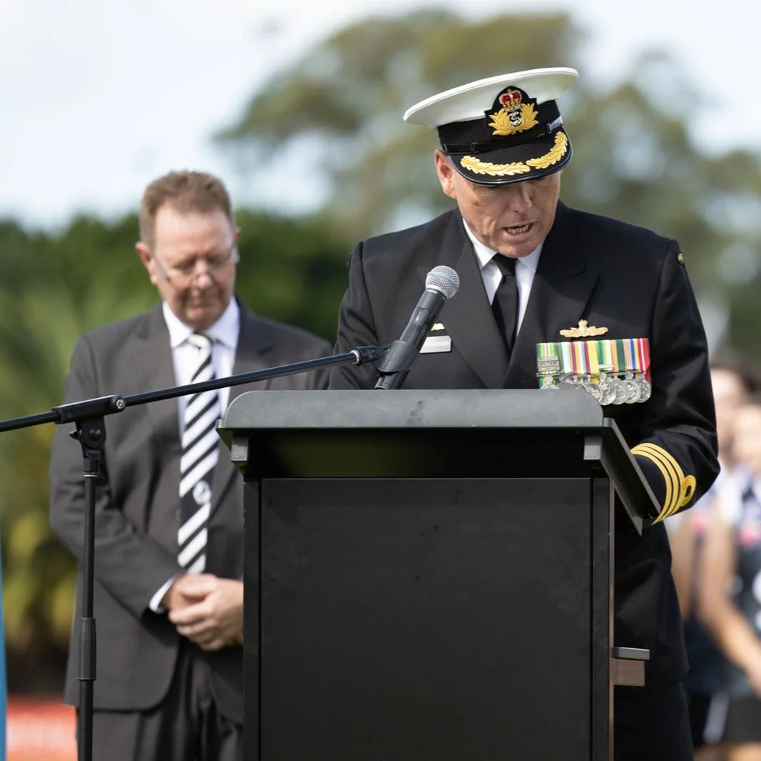 A military officer in uniform with medals giving a speech at a podium, with a man in a suit and tie standing behind him, outdoors during a formal event.