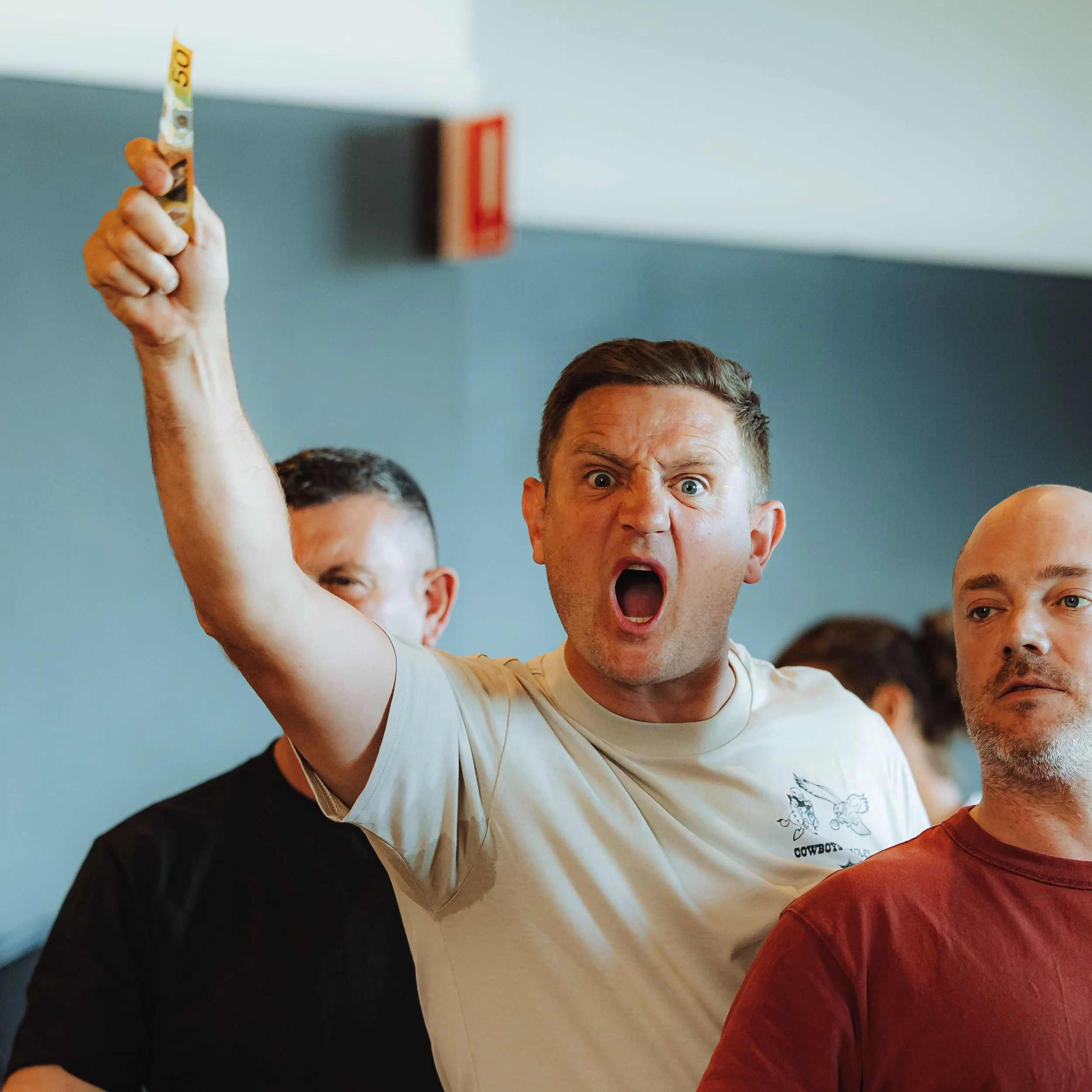 A man with short brown hair, wearing a beige T-shirt, is yelling and raising his hand holding a yellow object, among other men in a group.