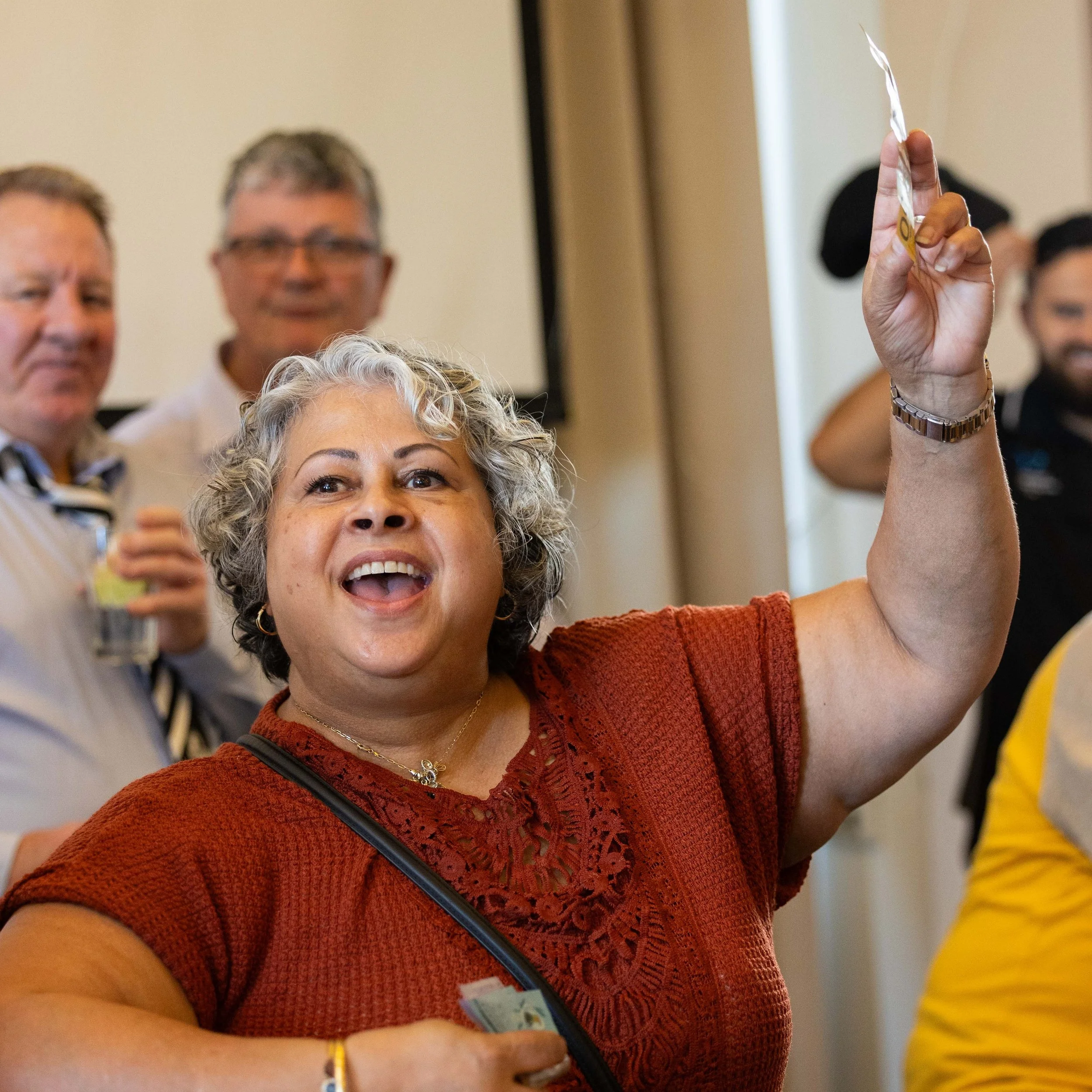 A woman with gray, curly hair smiling and holding up a small object in her right hand at a gathering or event, with several people in the background.