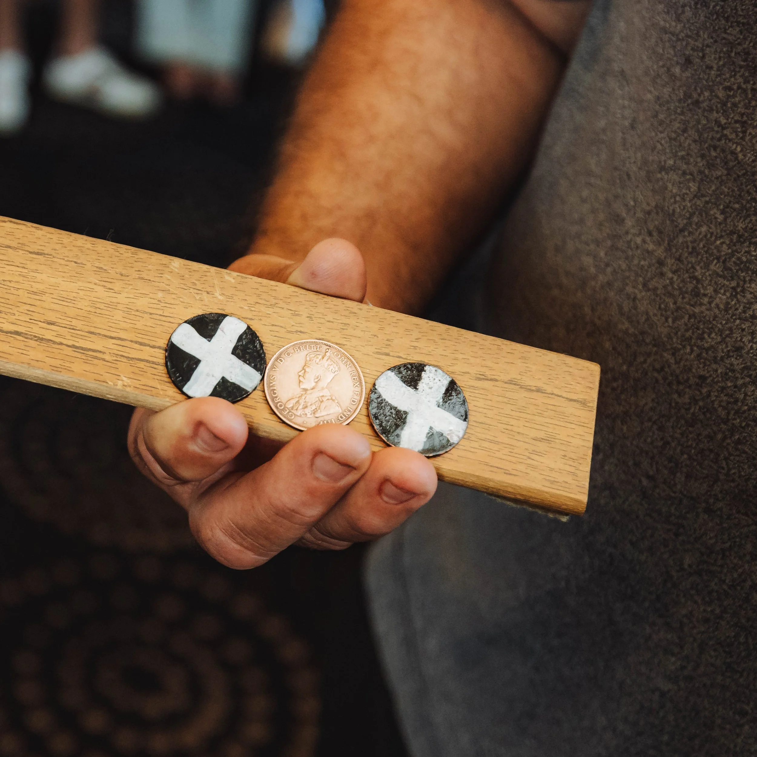 A person holding a wooden board with two buttons featuring black and white cross designs and a copper coin between them.