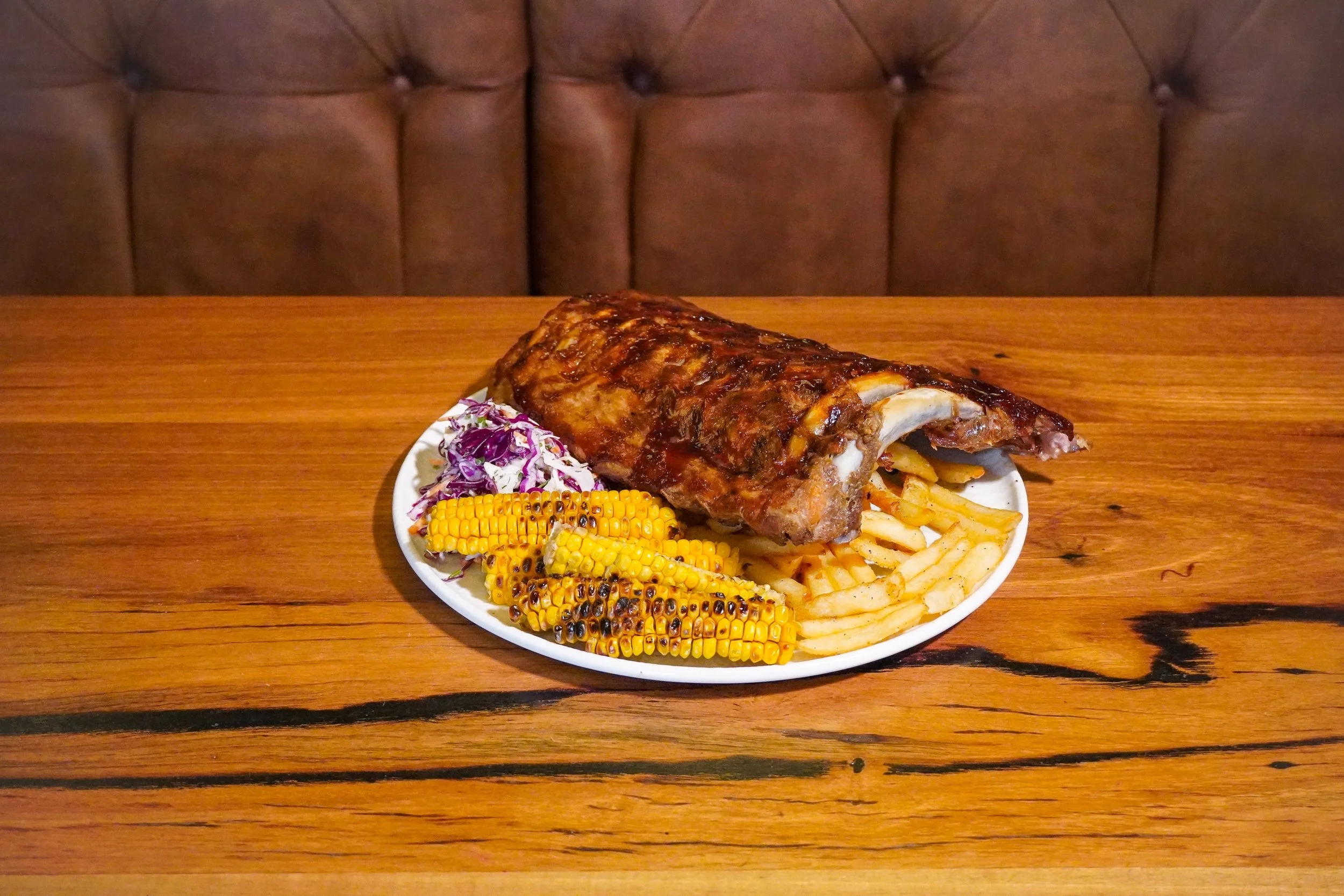 A plate of barbecue ribs with grilled corn, macaroni and cheese, and coleslaw on a wooden table in front of a tufted brown booth.