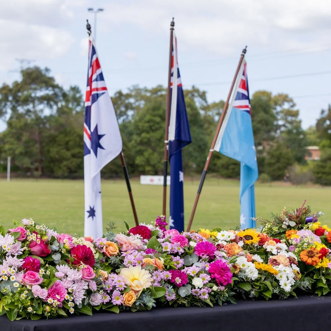 Three Australian flags on flagpoles with a background of green trees and cloudy sky, decorated with a colorful floral arrangement in the foreground.