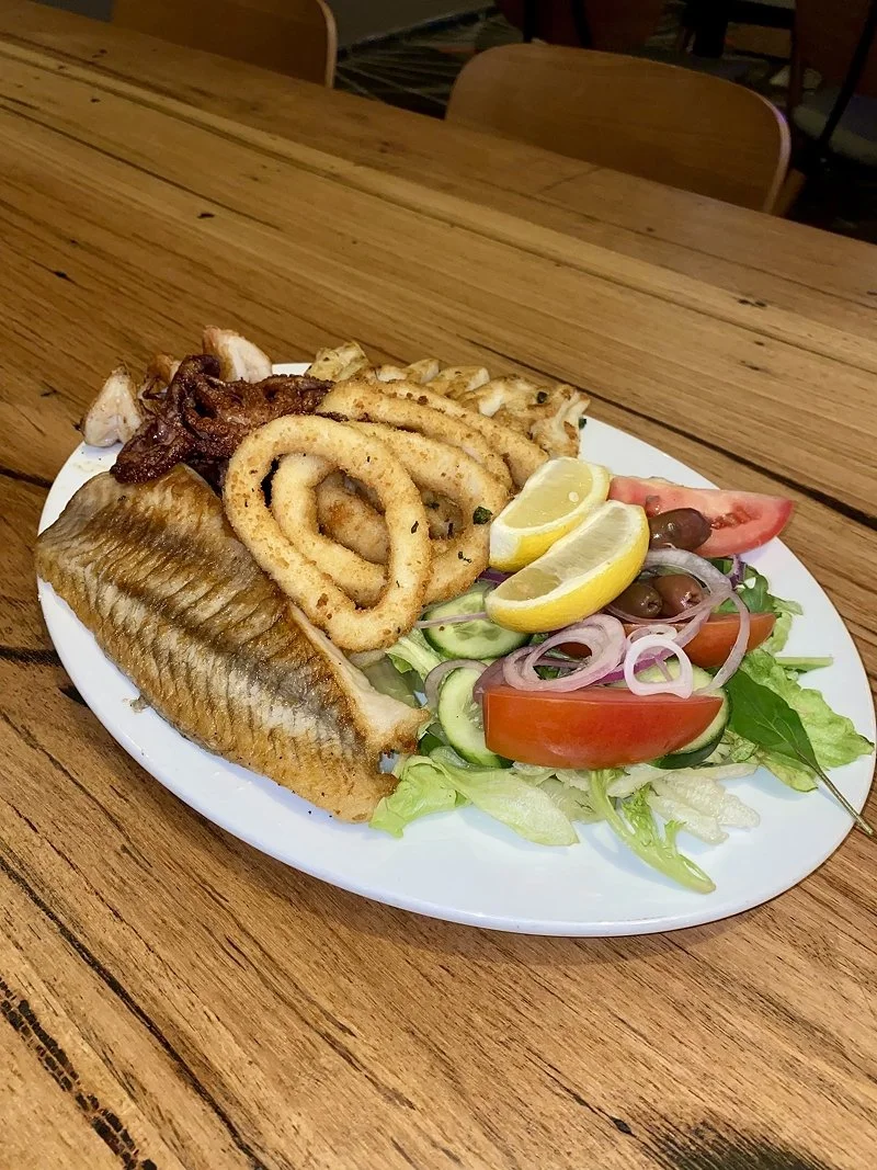 A plate of fried fish, fried calamari, onion rings, bacon, lemon wedges, and a salad with tomatoes, cucumbers, lettuce, and black olives on a wooden table.