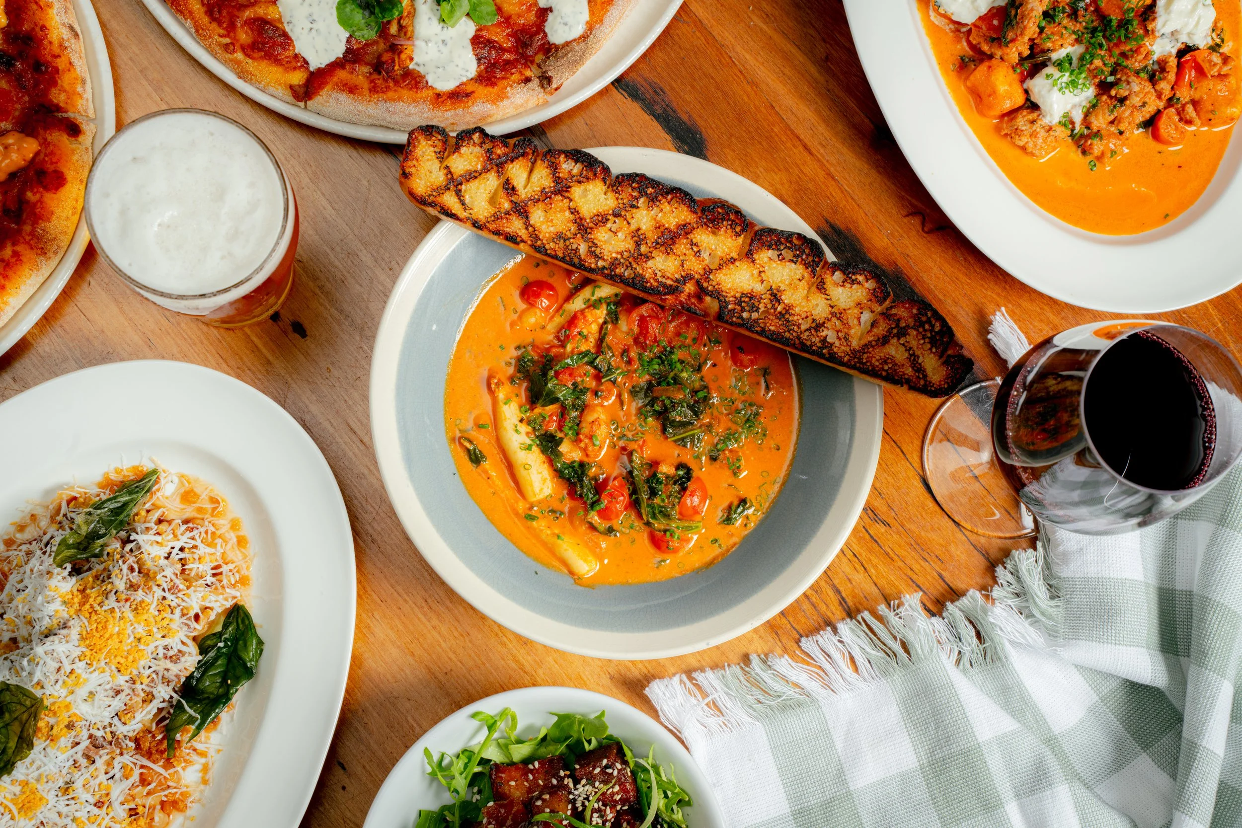A variety of Indian dishes on a wooden table, including a bowl of butter chicken curry, a plate of rice, grilled naan bread, a glass of beer, and a glass of red wine.