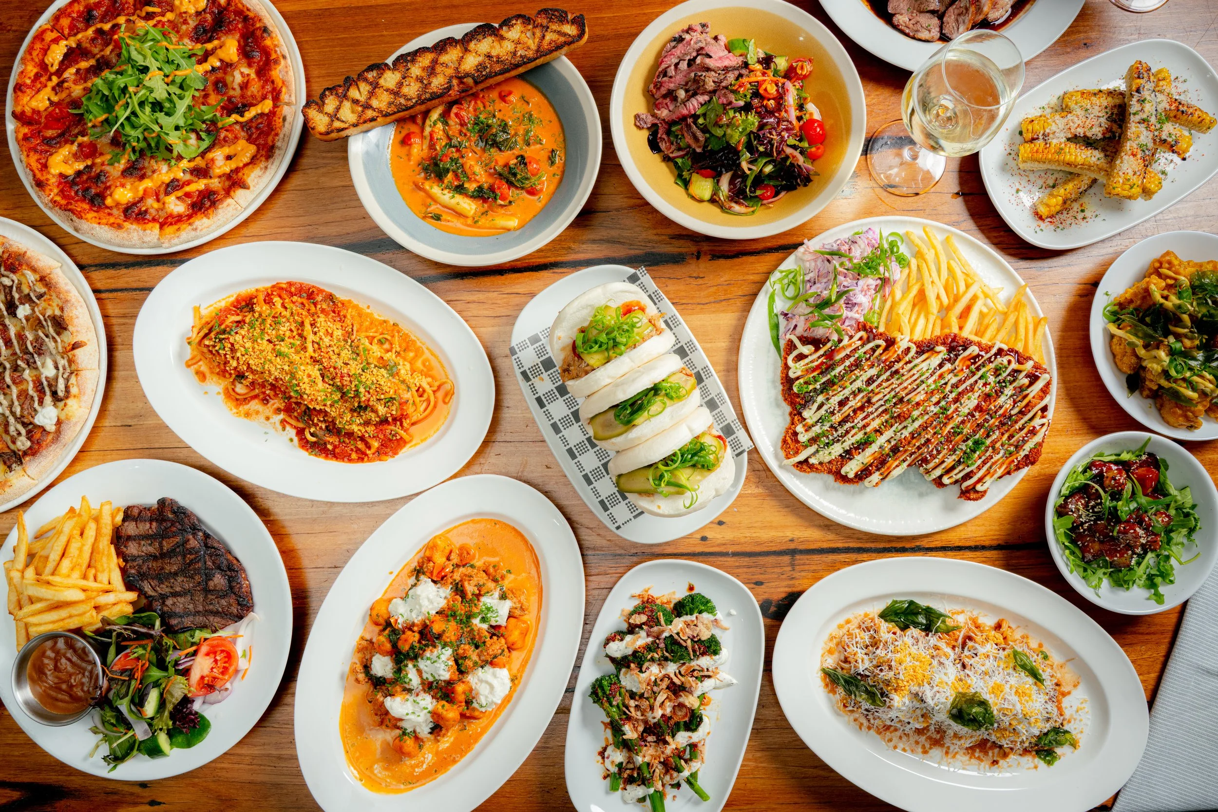 Assorted dishes of pizza, pasta, steak, salads, and appetizers on a wooden table.