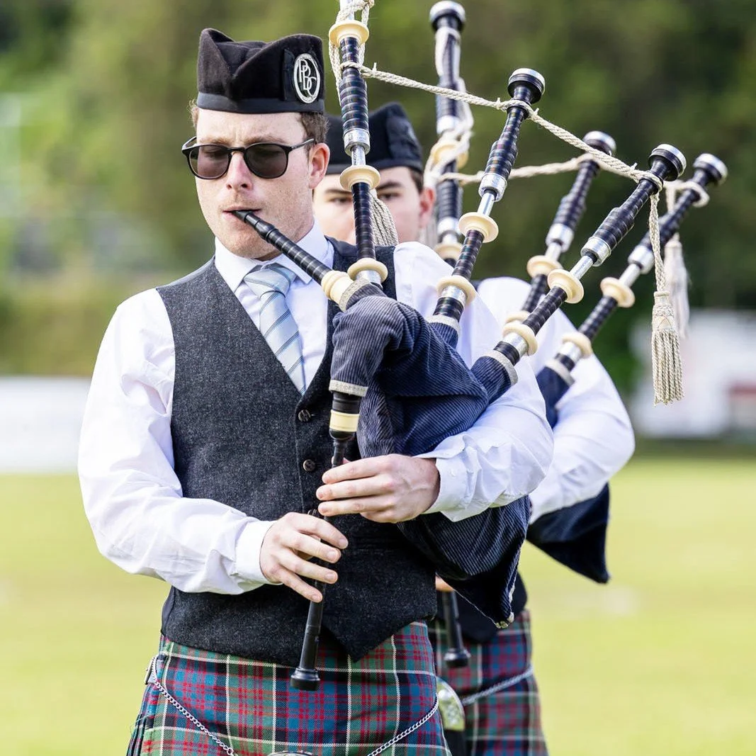 A man wearing traditional Scottish attire playing bagpipes outdoors, with another person partly visible in the background.