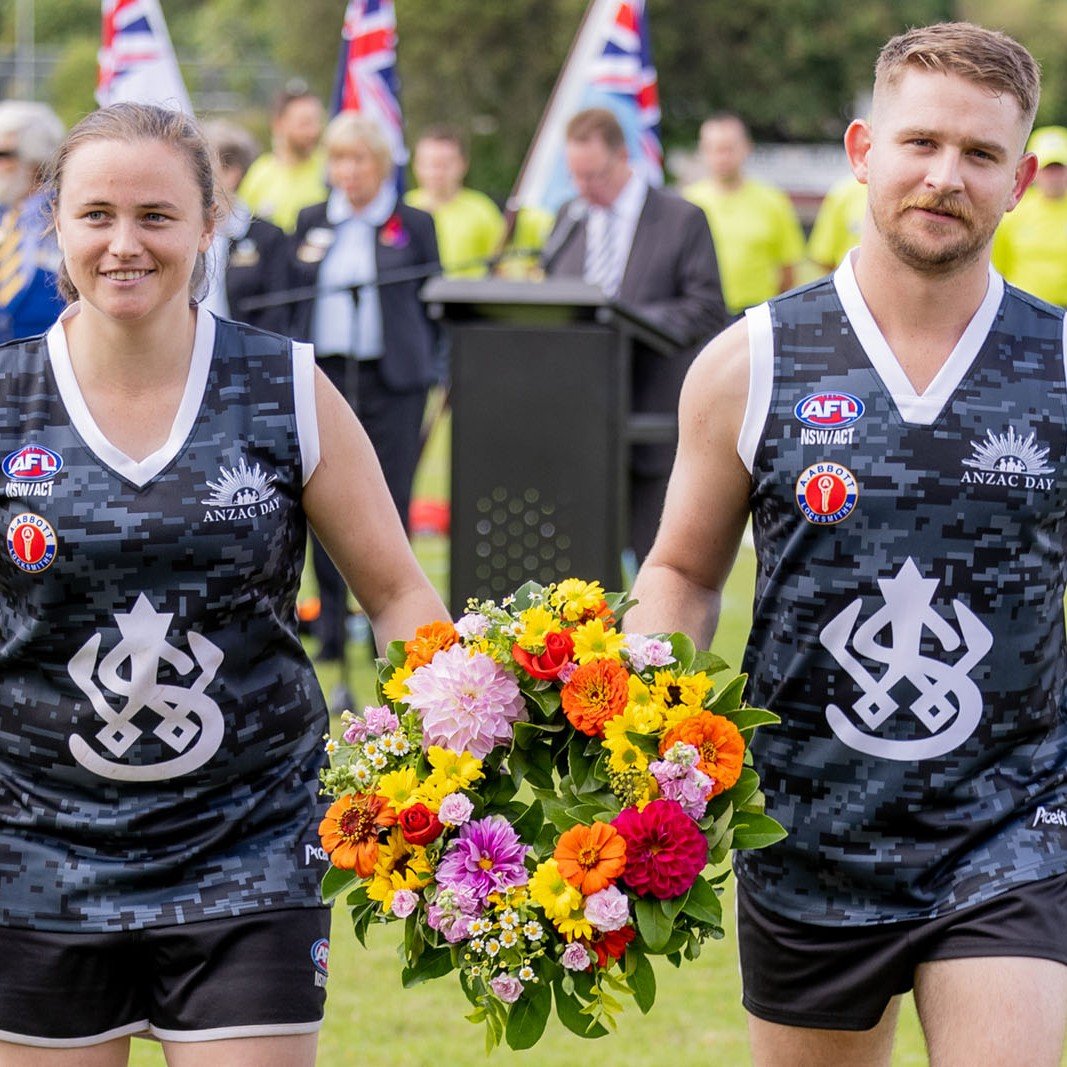 A woman and a man in Australian football jerseys holding a colorful flower wreath during a commemorative event at ANZAC Day, with onlookers and flag bearers in the background.