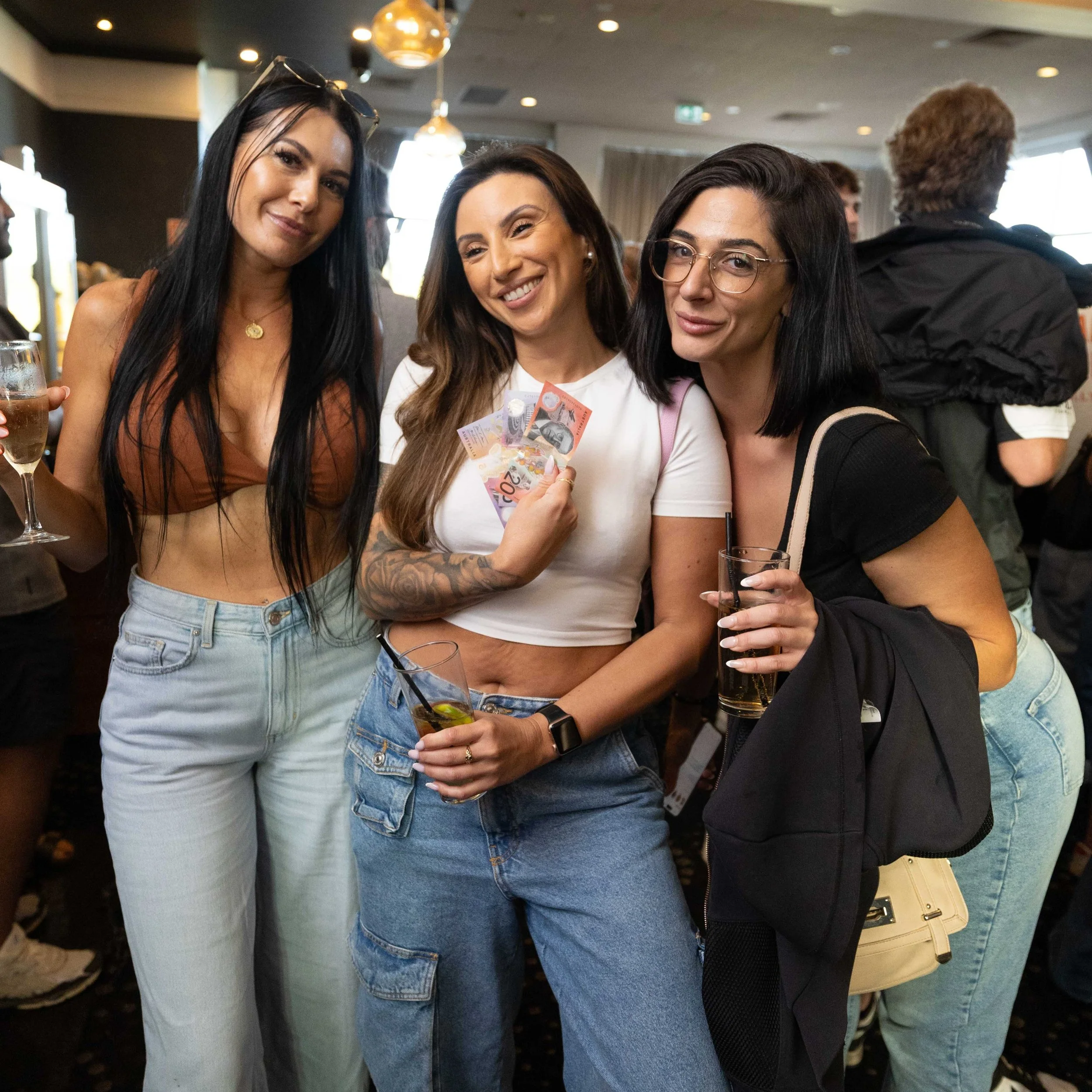 Three women smiling at a party, holding drinks, with one woman holding Australian currency notes.