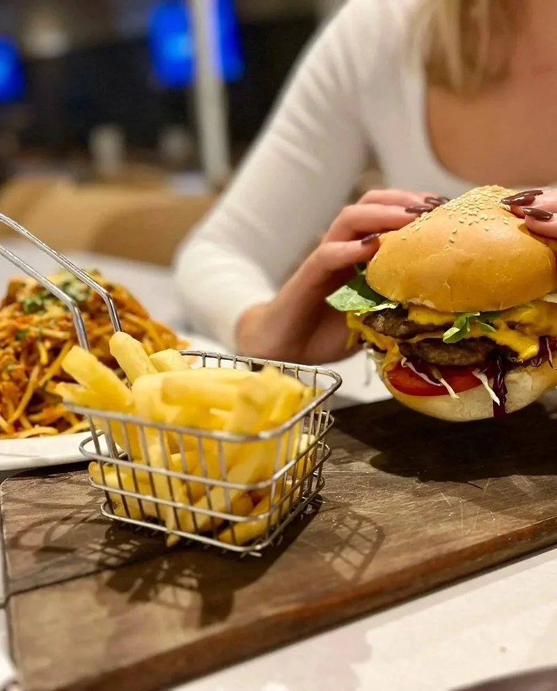 Close-up of a cheeseburger with lettuce, tomato, and cheese, held by a woman with long nails, alongside a small basket of French fries and a plate of pasta on a wooden table in a restaurant.