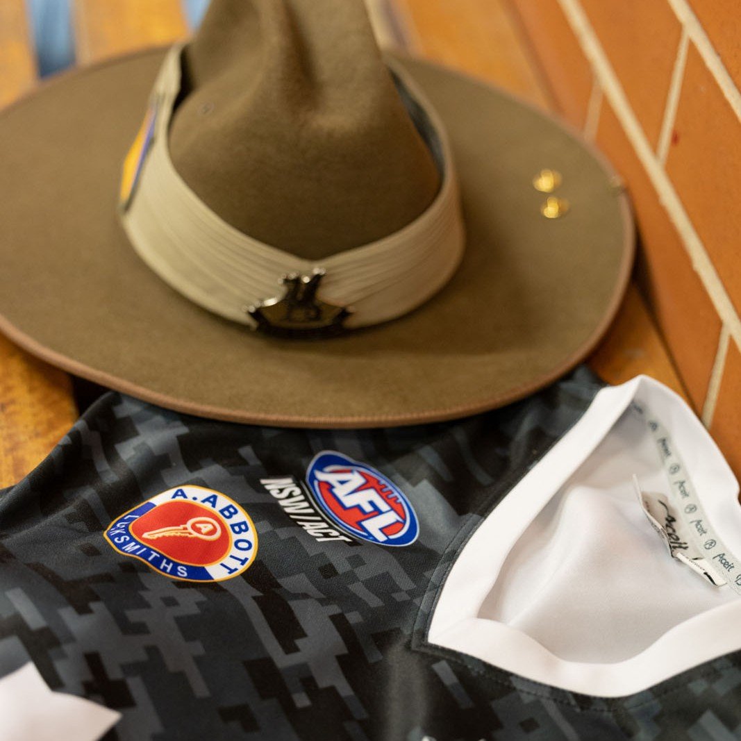A brown cowboy hat with a black badge, a black sports jersey with logos including AFL, AFLW, and Shultz Auto, and a brick wall in the background.