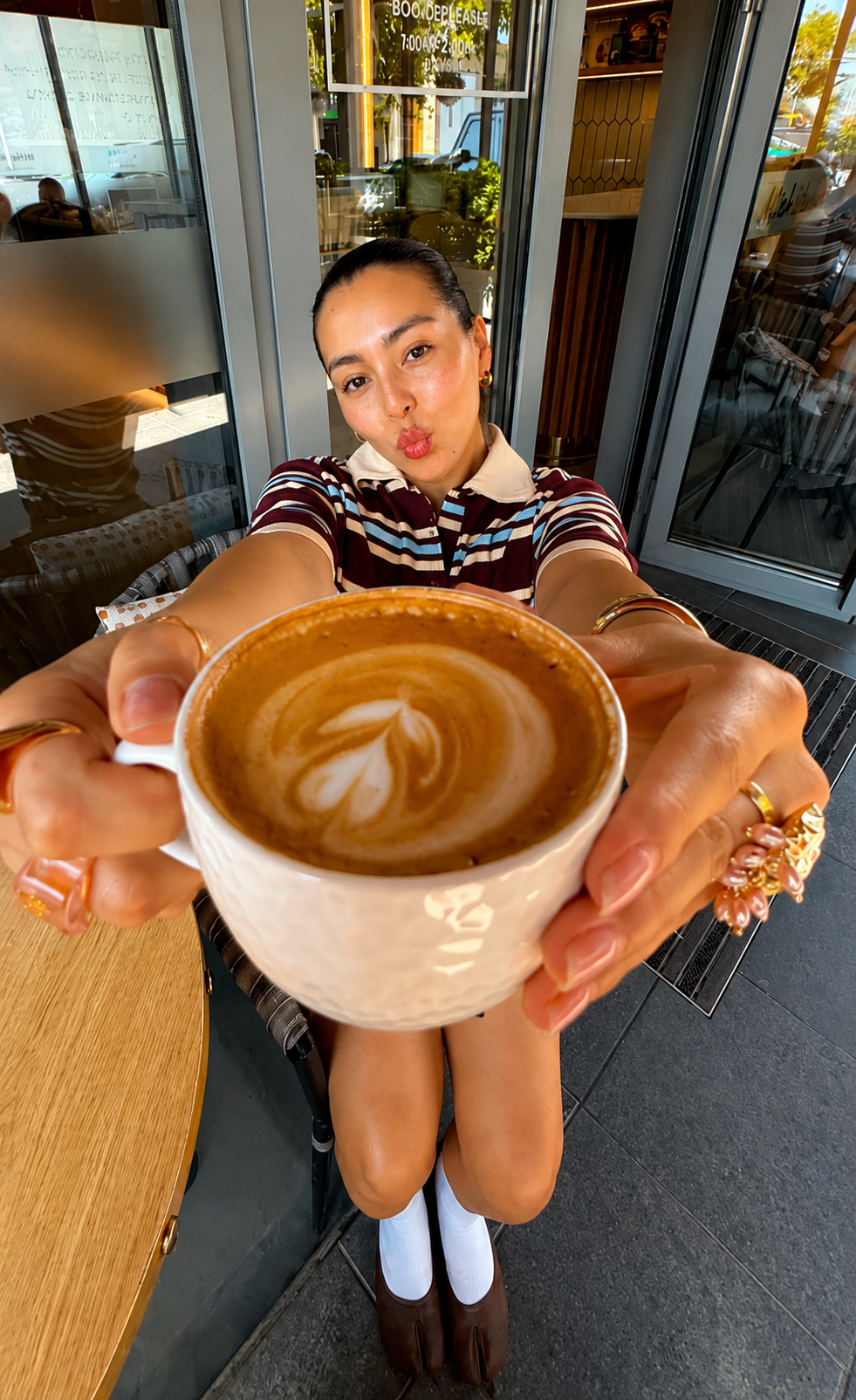 A woman sitting in a cafe holding a white ceramic mug filled with coffee featuring latte art. She is making a kissy face towards the camera, wearing a striped shirt, jewelry, and white socks with brown shoes. The cafe has large windows with sunlight and outdoor seating visible.