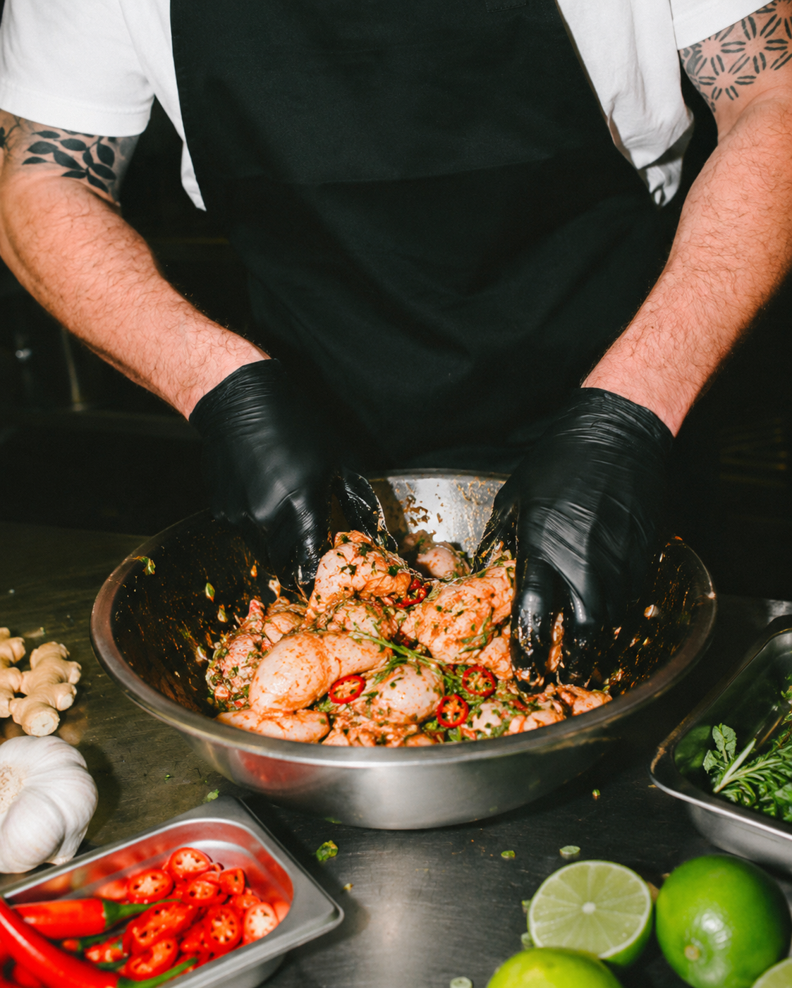 Person wearing black gloves mixing raw chicken pieces with spices in a stainless steel bowl, surrounded by garlic, red chili peppers, lime, and herbs on a dark tabletop.