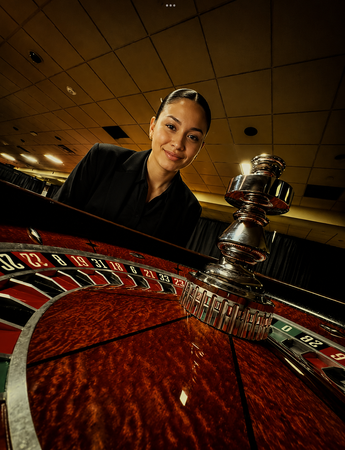 A woman with dark hair and a black blazer smiling near a roulette wheel in a casino.