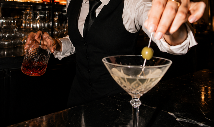 A bartender in a black vest and white shirt preparing a cocktail with an olive on a skewer in front of a dark bar background.