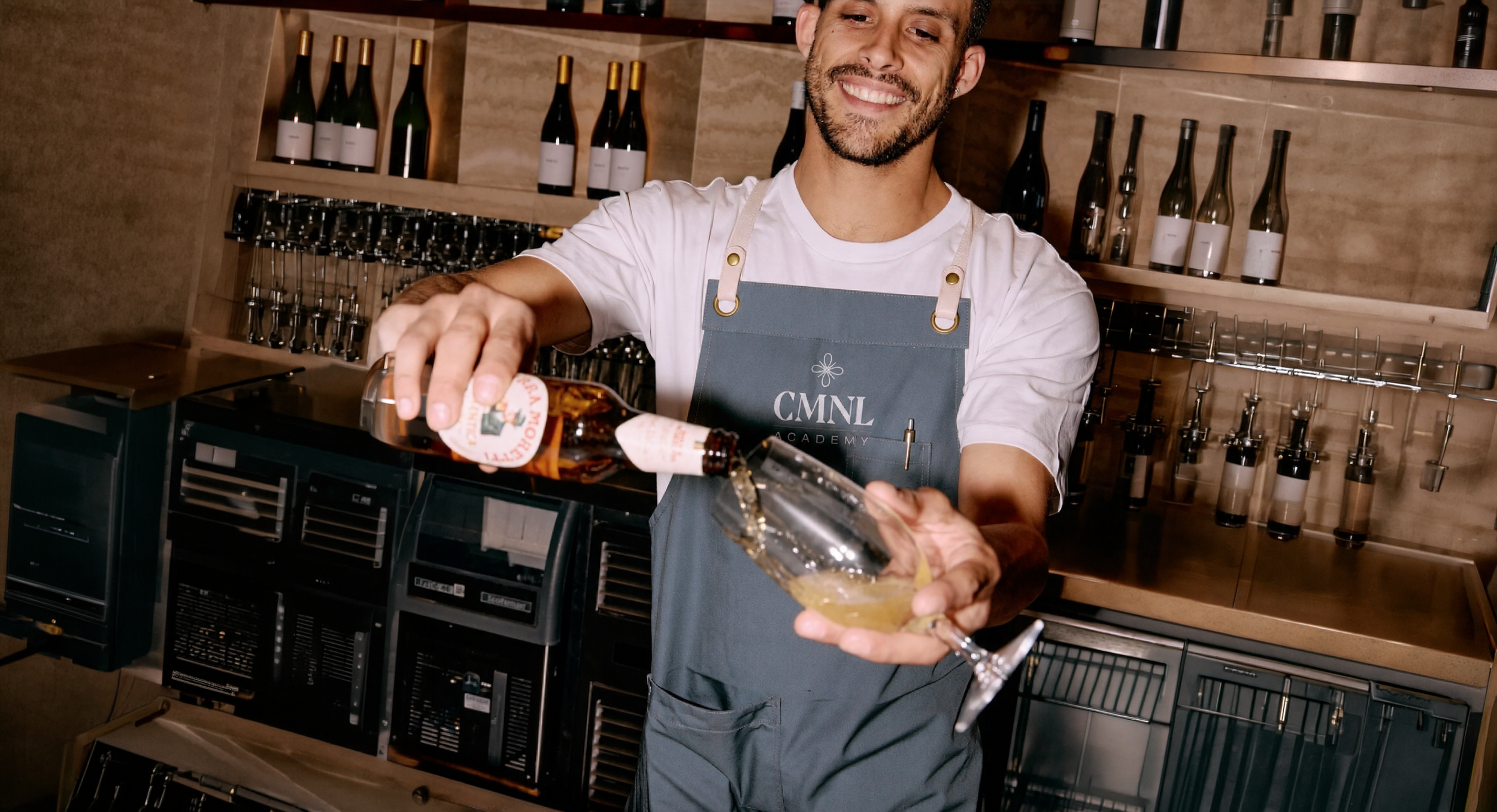 A smiling man wearing a gray apron is pouring a drink, likely champagne or sparkling wine, into a coupe glass in a bar or restaurant setting with bottles and wine dispensers in the background.